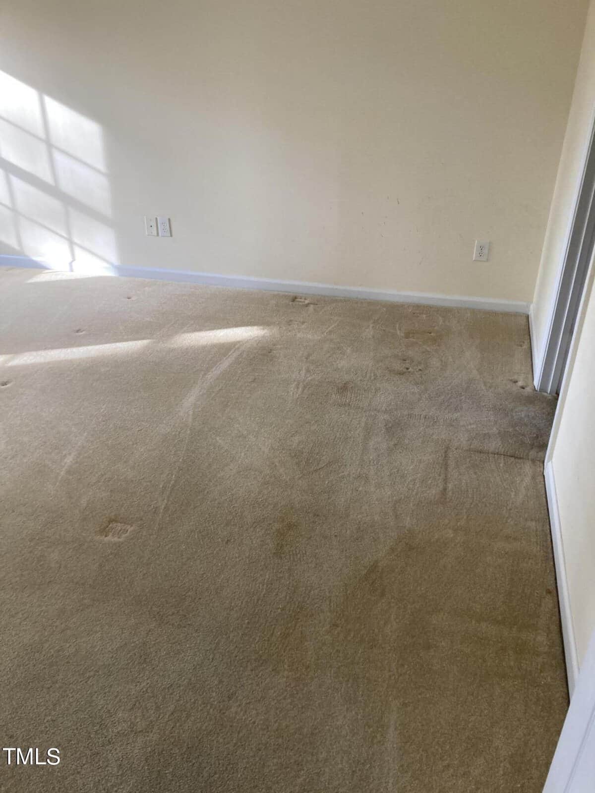 A room with light-colored carpet showing some stains and wear. Sunlight streams in from a window, casting grid-like shadows on the wall. The walls are painted light beige, and there are electrical outlets visible.