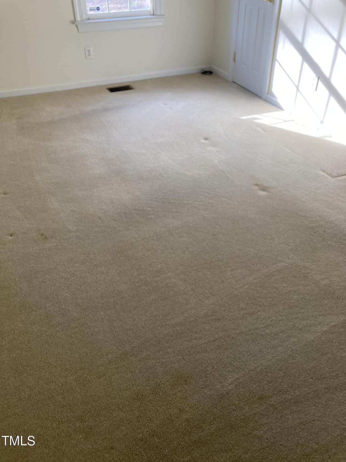 A sunlit, empty room with beige carpet, a white wall, and a window partially visible on the left. Sunlight casts a soft glow through glass doors on the right, illuminating the floor.