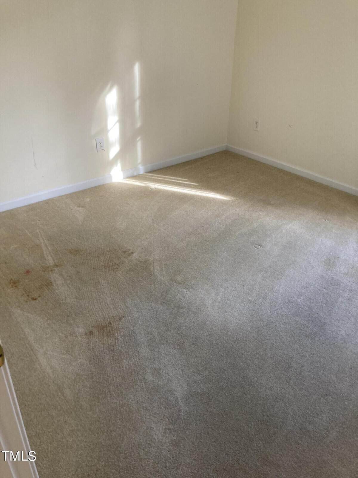 An empty room with beige carpet showing some stains and signs of wear. The walls are light-colored, and sunlight is filtering in, creating a pattern on the carpet. The room appears to have minimal furnishings.
