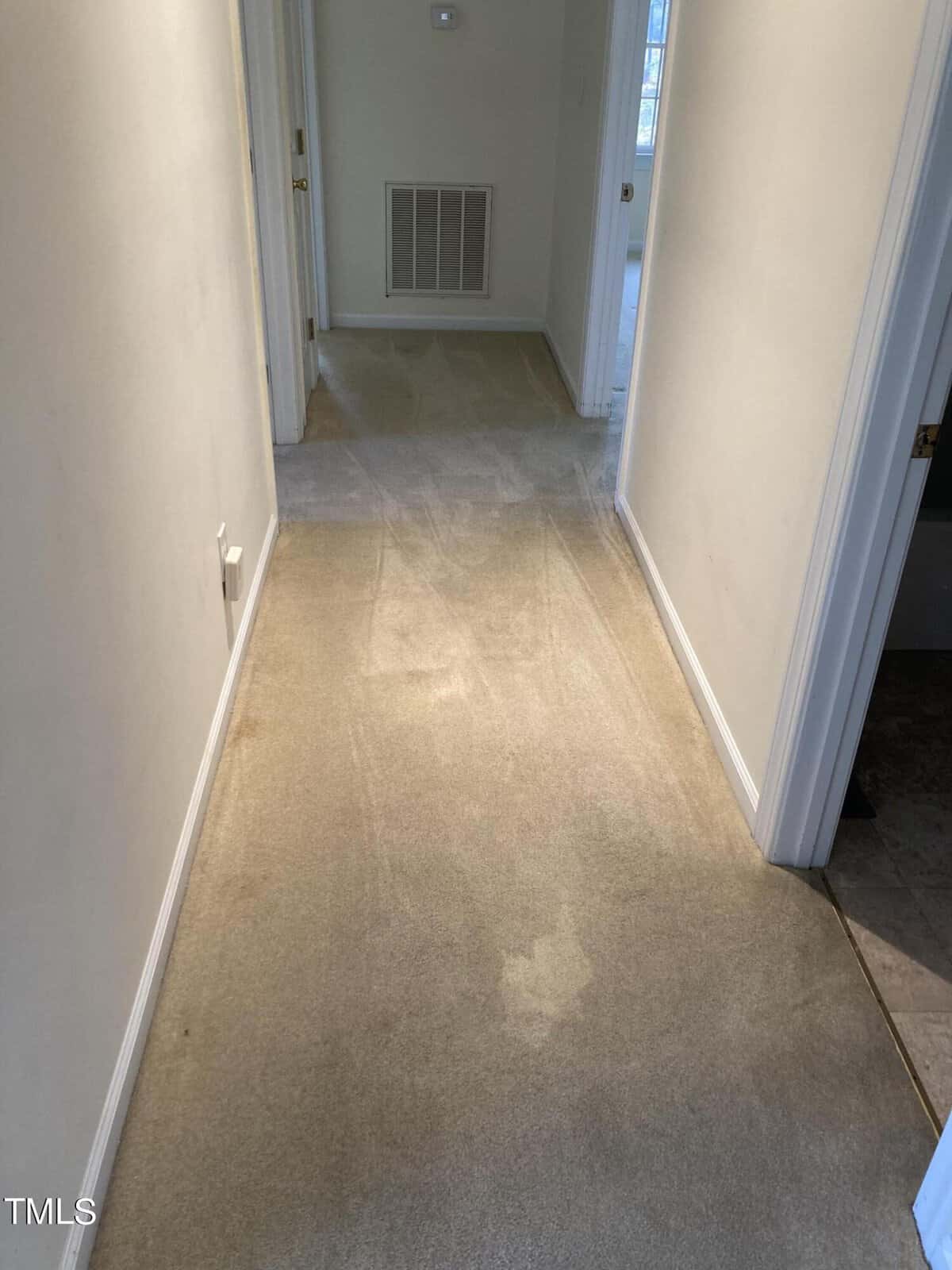 A clean, beige carpeted hallway with off-white walls and white trim. There are doorways on both sides leading to different rooms. The hallway is well-lit by natural light from nearby windows.