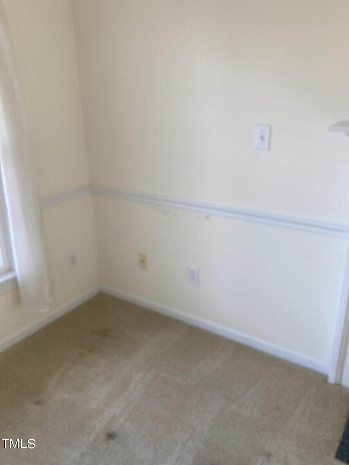 A corner of a room with beige carpeting and light yellow walls. The walls have a white chair rail molding, electrical outlets, and a light switch. A window with sheer curtains is partially visible on the left side.