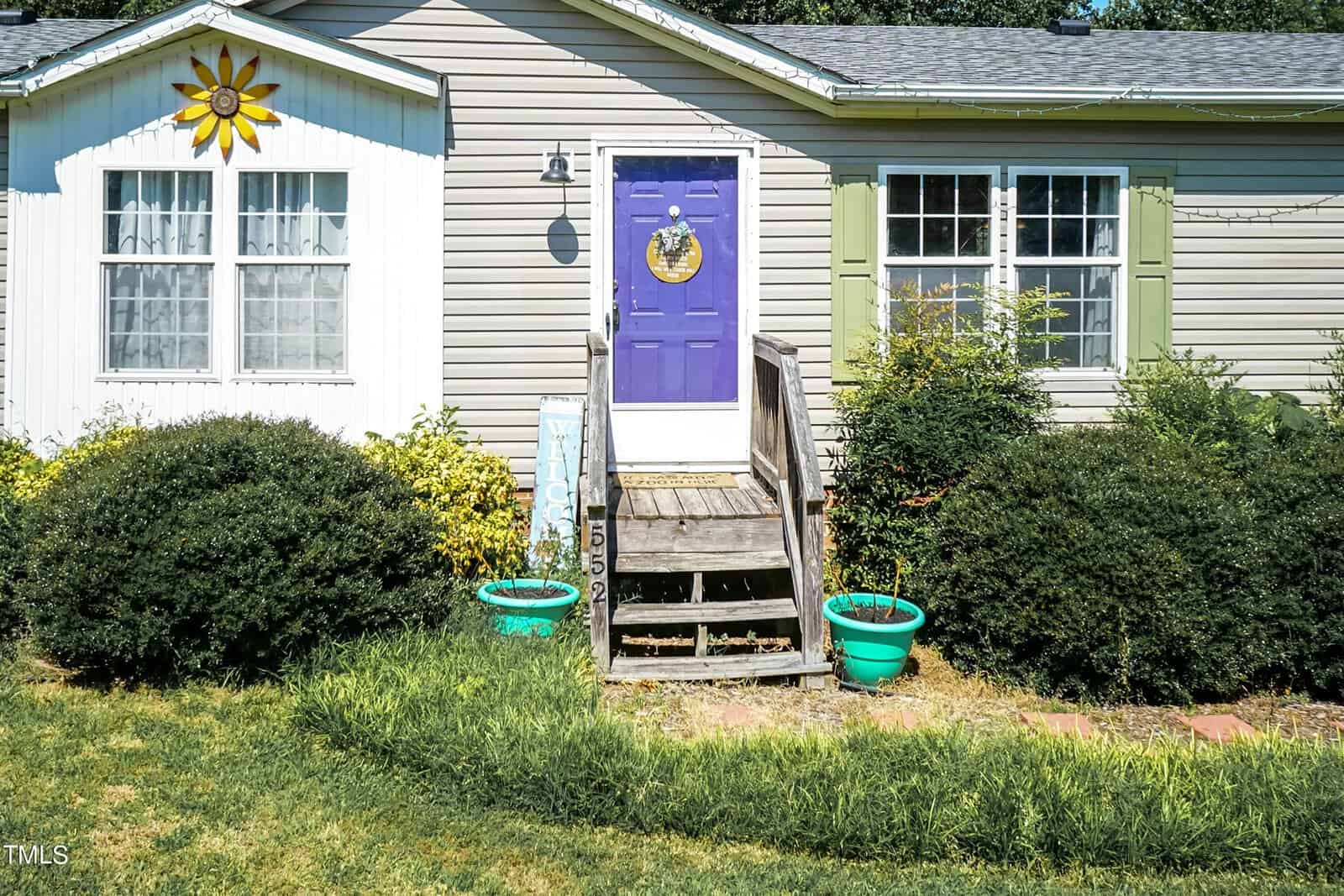 A charming small house on 552 Locks Lane features light gray siding and a distinctive purple front door with a sun ornament above its window. Two green planters grace the steps, displaying its number proudly. Nestled in Rougemont, the house is surrounded by lush bushes and greenery.