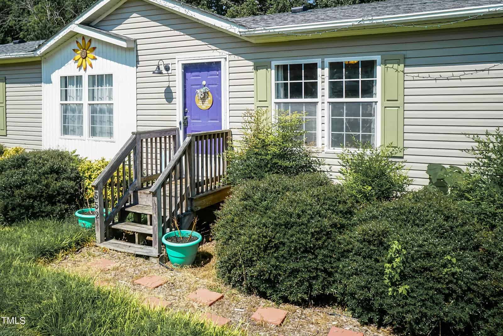A small house with green and white siding and a purple door featuring a wreath. A yellow sun decoration is above the front windows. There are bushes and two turquoise planters beside the short wooden steps leading to the entrance.
