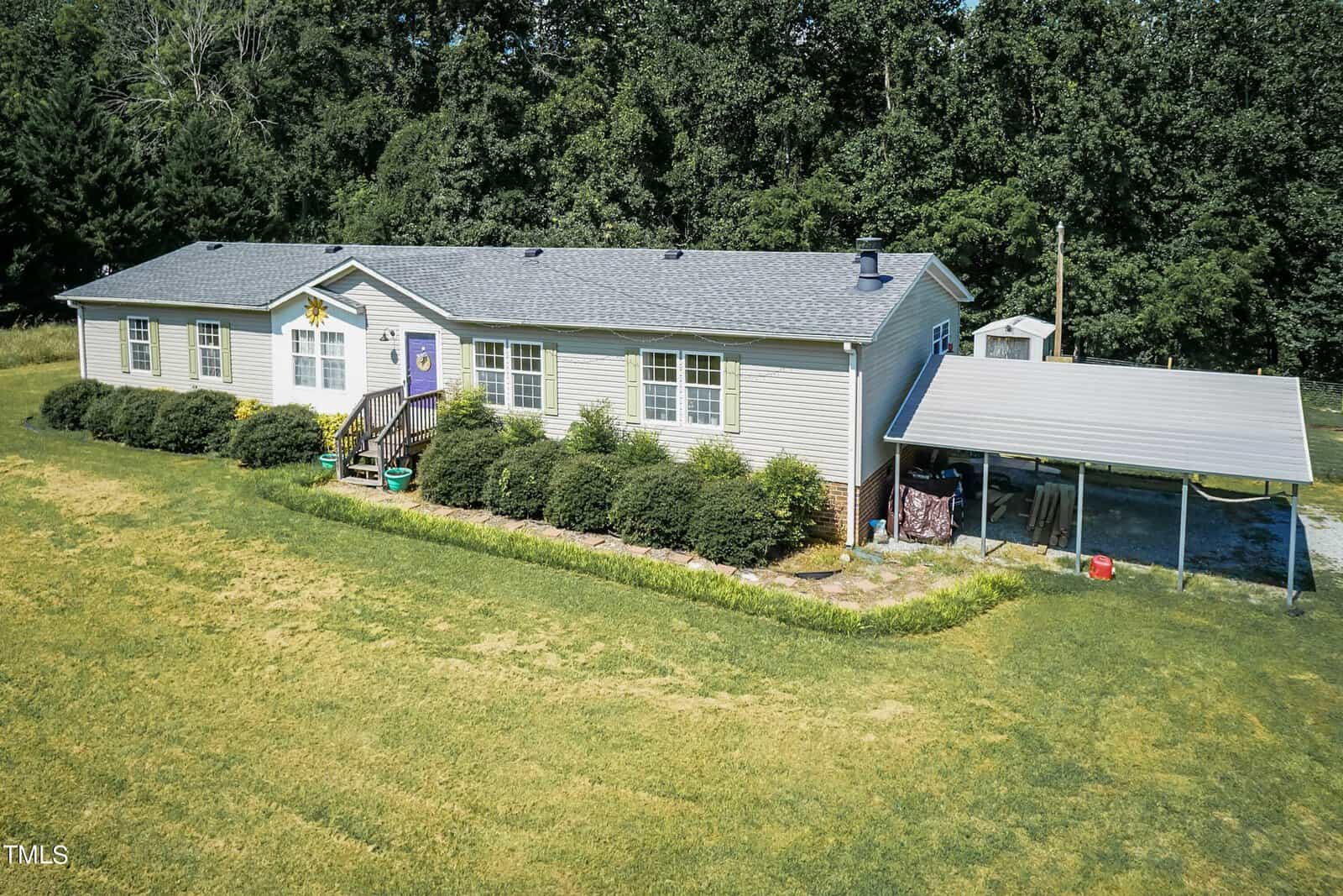 Aerial view of a single-story gray house with a pitched roof. It features a front porch and a carport on the right side. The house is surrounded by neatly trimmed bushes, and a large lawn extends in front, bordered by dense trees in the background.