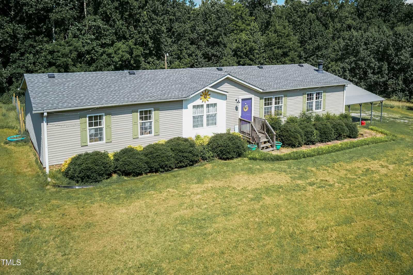 A single-story house with a gray roof and beige siding sits in a grassy yard. The front features a small porch with steps, a purple door, and windows with white trim. Lush shrubs line the front, and a wooded area is visible in the background.