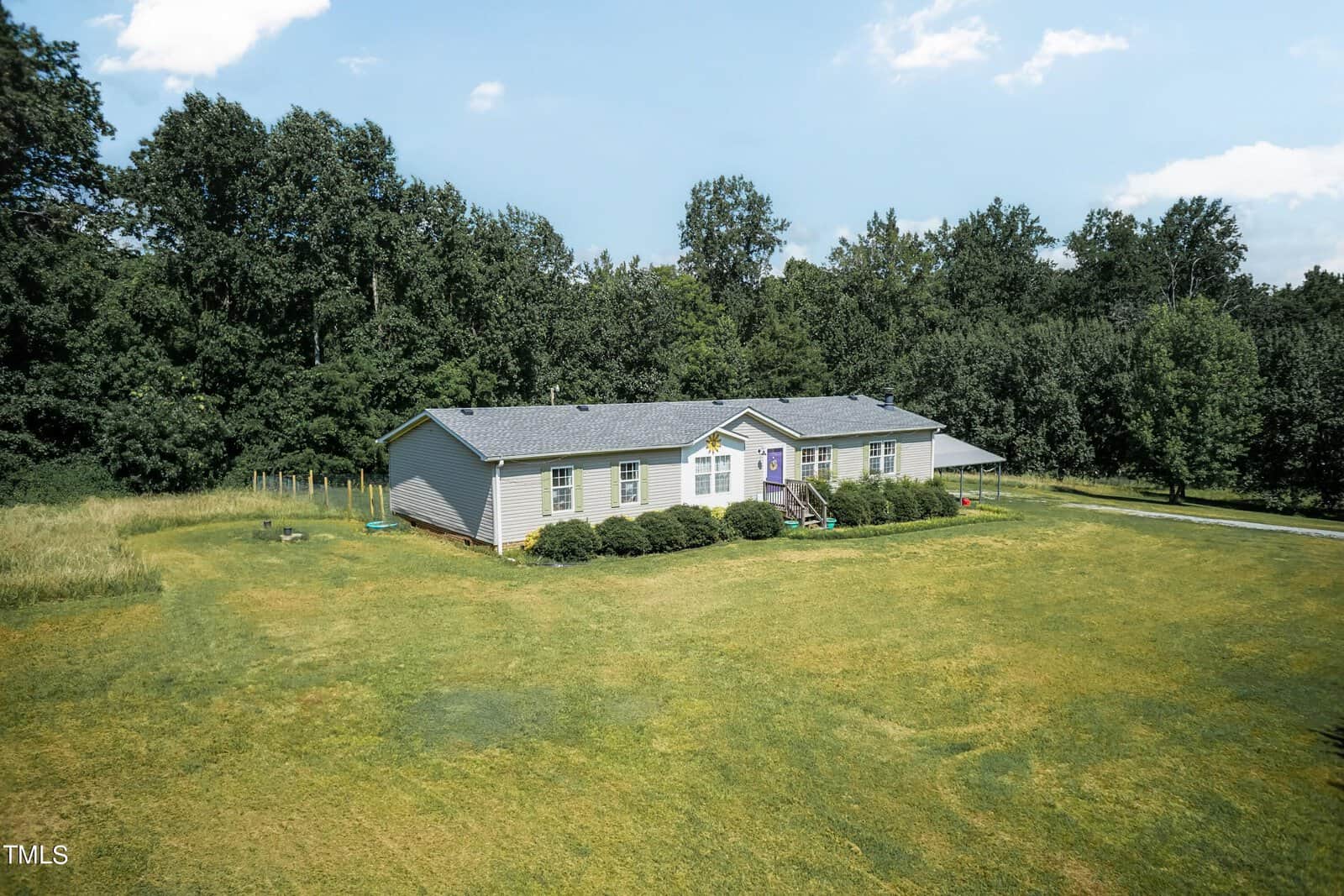 Aerial view of a single-story house with a gray roof, surrounded by a large grassy yard and bordered by dense woodland. The sky is partly cloudy.