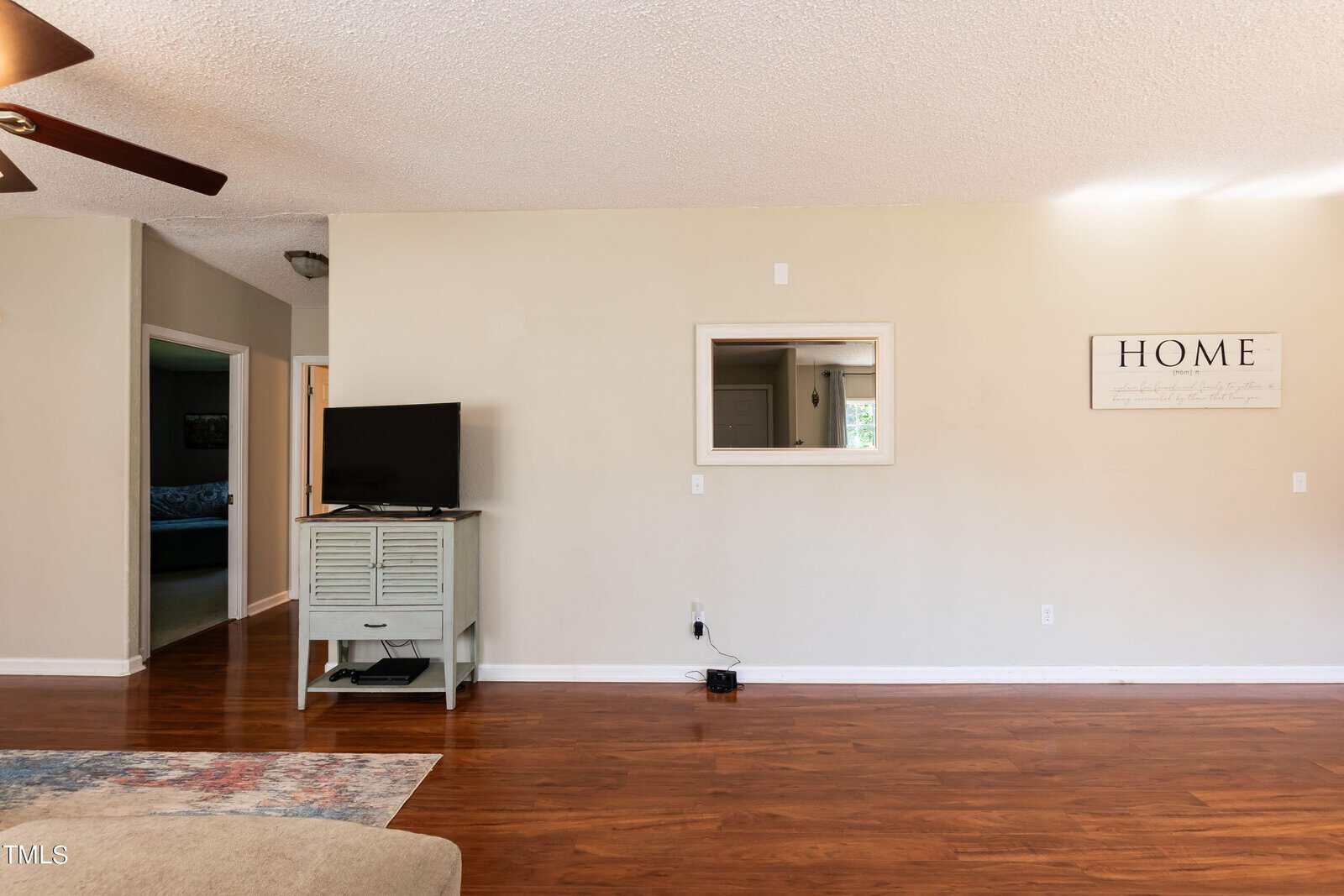 A living room with a wooden floor, beige walls, a light blue TV stand with a television, a mirror, and a wall sign that says "Home." A patterned rug and part of a beige couch are visible in the foreground. Ceiling fan blades are also visible.