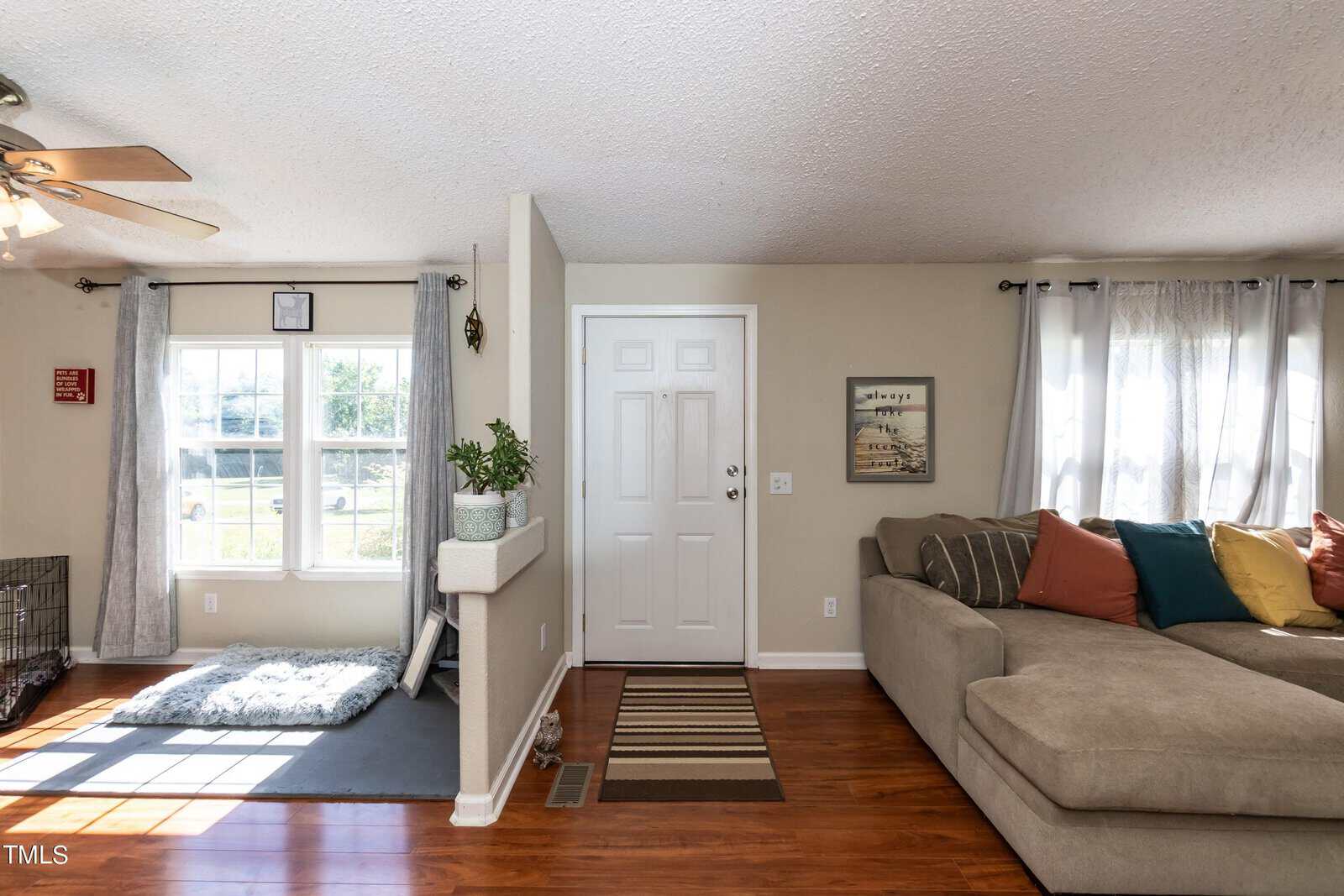 A cozy living room with a large beige sectional sofa adorned with colorful cushions. Sunlight streams in through a window with white curtains. A small plant sits on a shelf near the white front door, and a striped rug lies on the wooden floor.