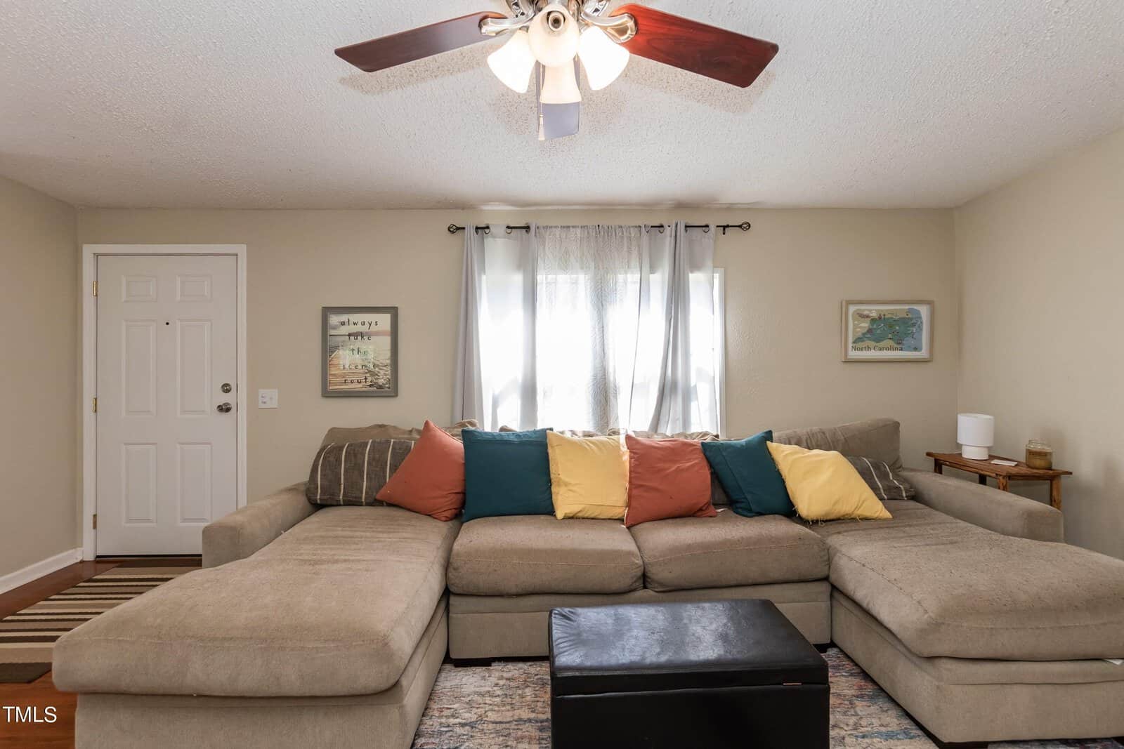 A cozy living room with a large beige sectional sofa adorned with colorful pillows. A small black ottoman sits in front of the sofa. Natural light filters through sheer curtains on the window behind. A ceiling fan with lights hangs overhead.