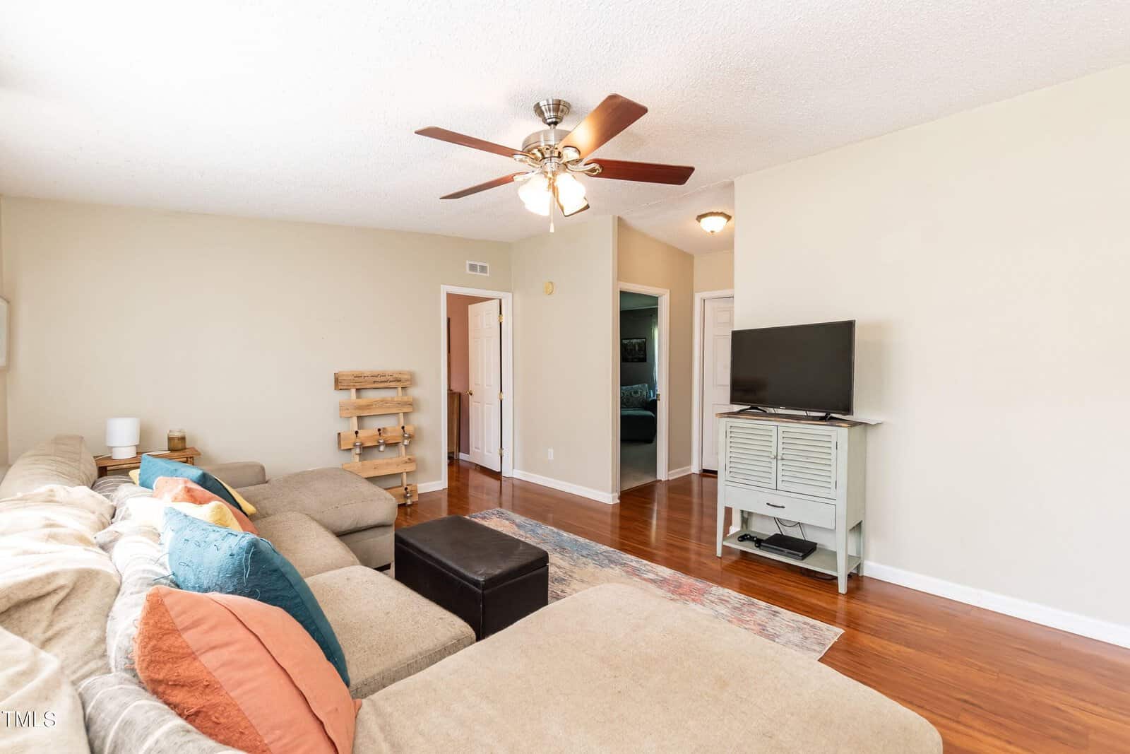 A cozy living room with a beige sectional sofa adorned with colorful pillows. A ceiling fan hangs overhead, and a TV sits on a console against a light-colored wall. Wooden floors and soft lighting create a warm atmosphere.