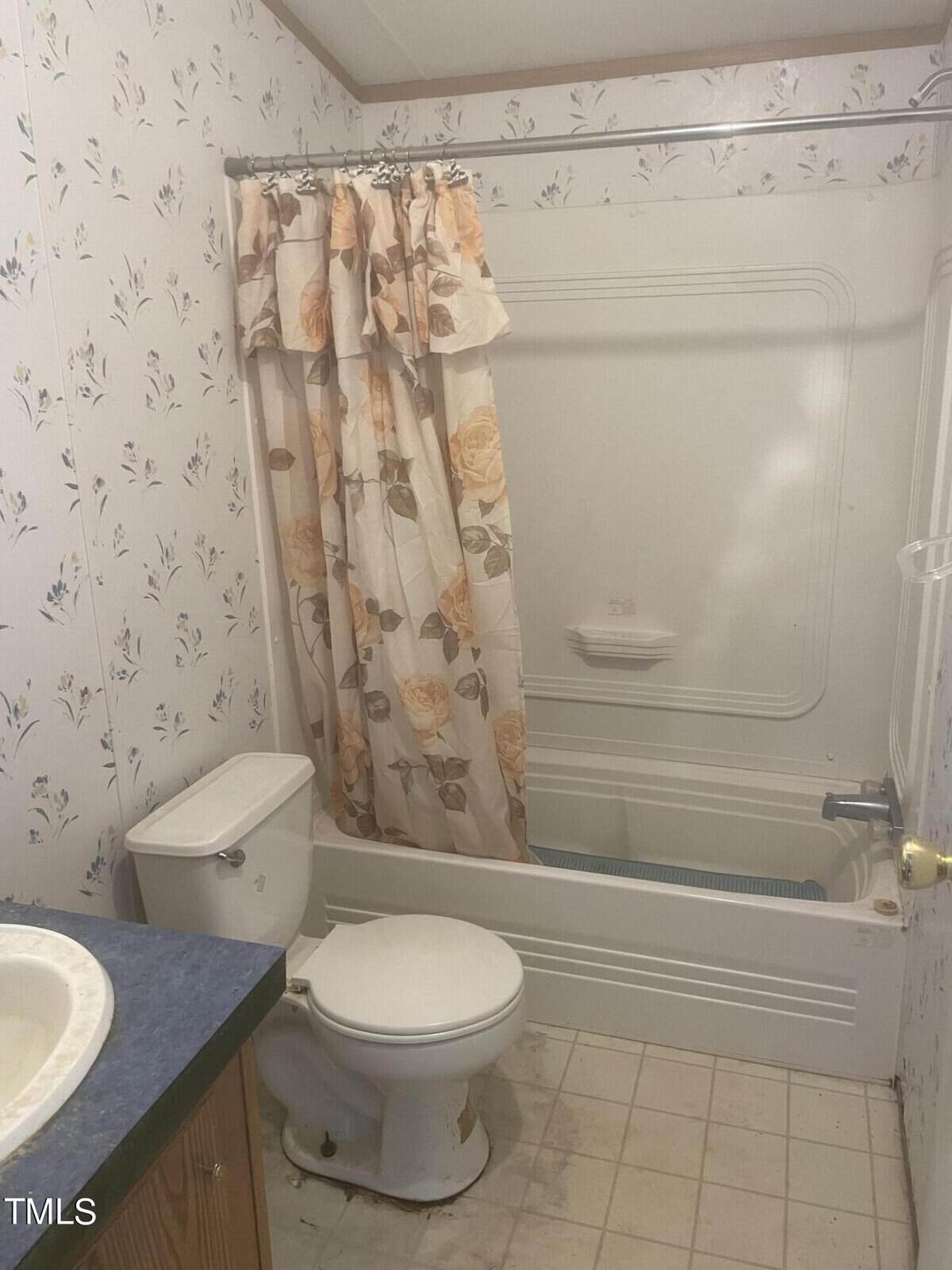 A small bathroom with floral wallpaper features a bathtub with a beige and peach floral shower curtain. There is a white toilet and a countertop with a sink on the left side. The floor is tiled, and the bathroom has a modest, vintage appearance.