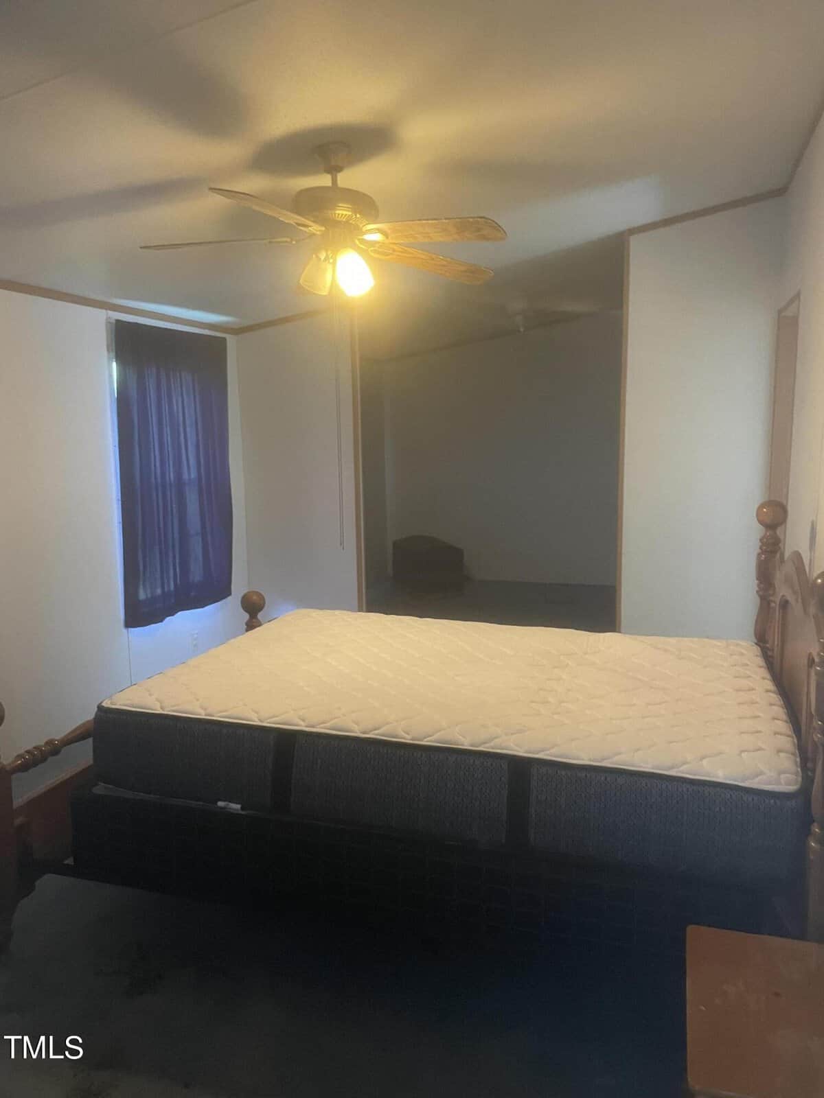 A dimly lit bedroom with a ceiling fan and a window covered by a dark curtain. The room features a wooden bed frame with a mattress placed on it, and a small wooden nightstand beside the bed.
