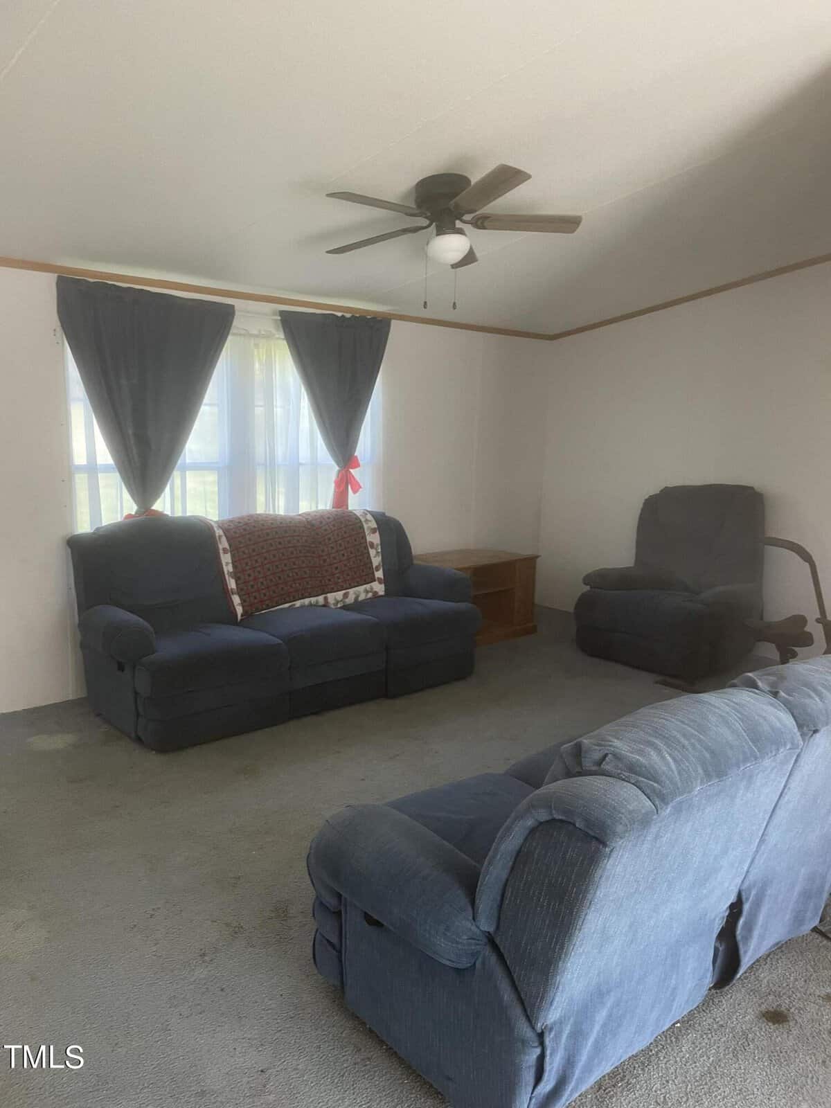 A living room with two blue sofas facing each other, a blue recliner, and a ceiling fan above. There's a window with dark curtains and a quilt draped over one sofa's back. The carpet is gray, and a small wooden table is near the window.