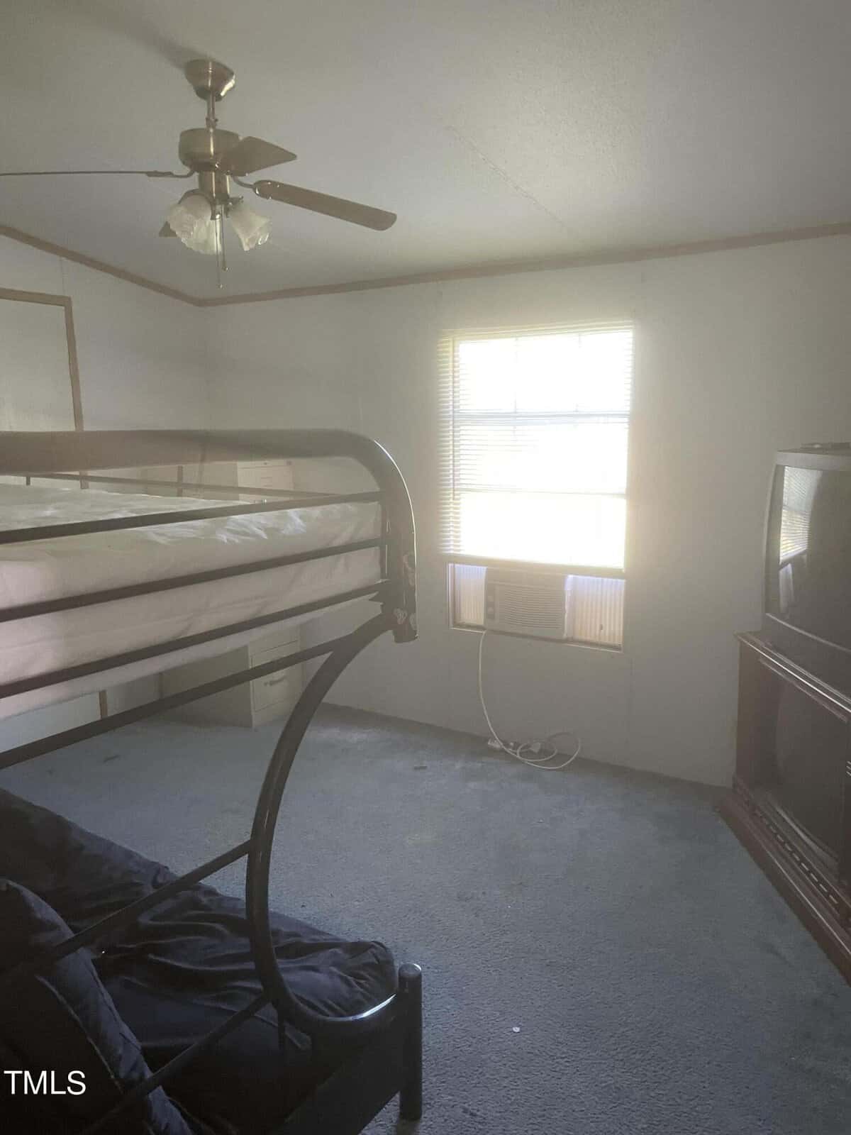 A bedroom with a metal bunk bed, a window with an air conditioner, and a TV on a stand. The room has a carpeted floor, a ceiling fan, and light coming in from the window.