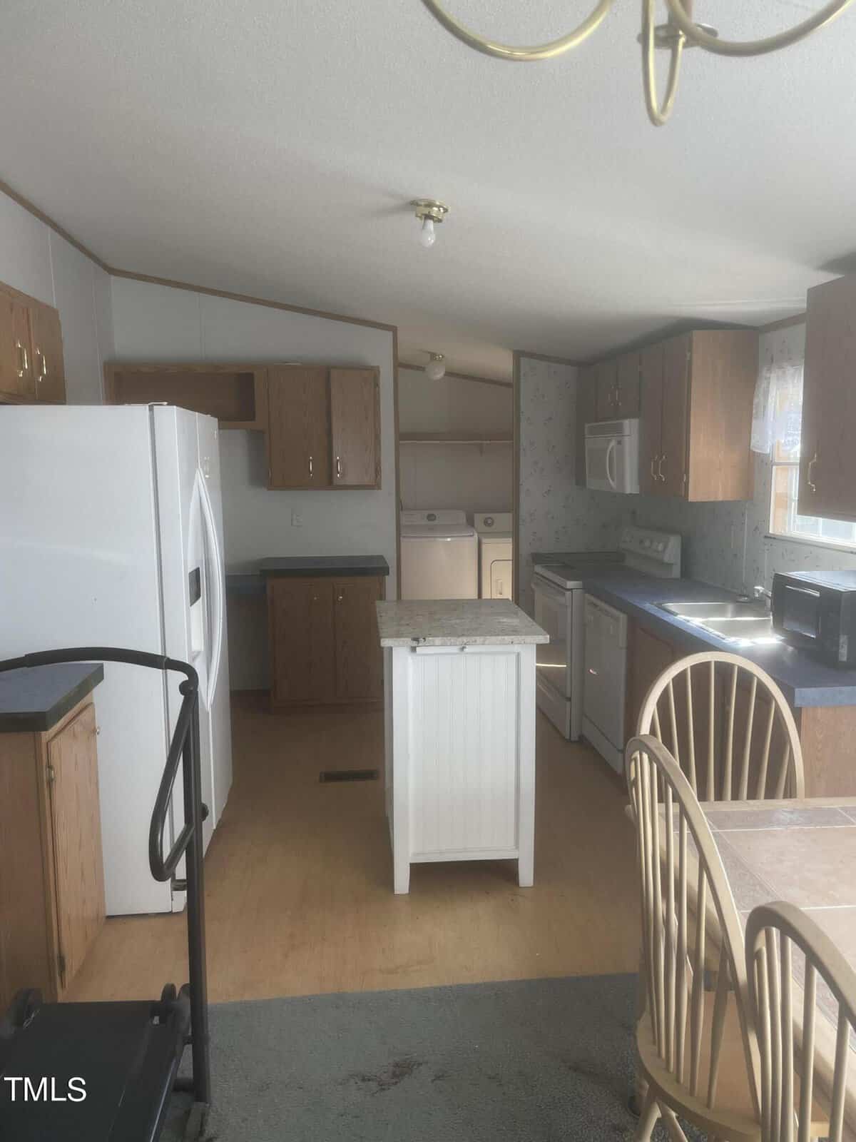 A cozy kitchen with wooden cabinets, a white refrigerator, and a small island with a granite top. There is a stove, microwave, and dishwasher. A table with wooden chairs is visible, and a treadmill is placed near the counter.