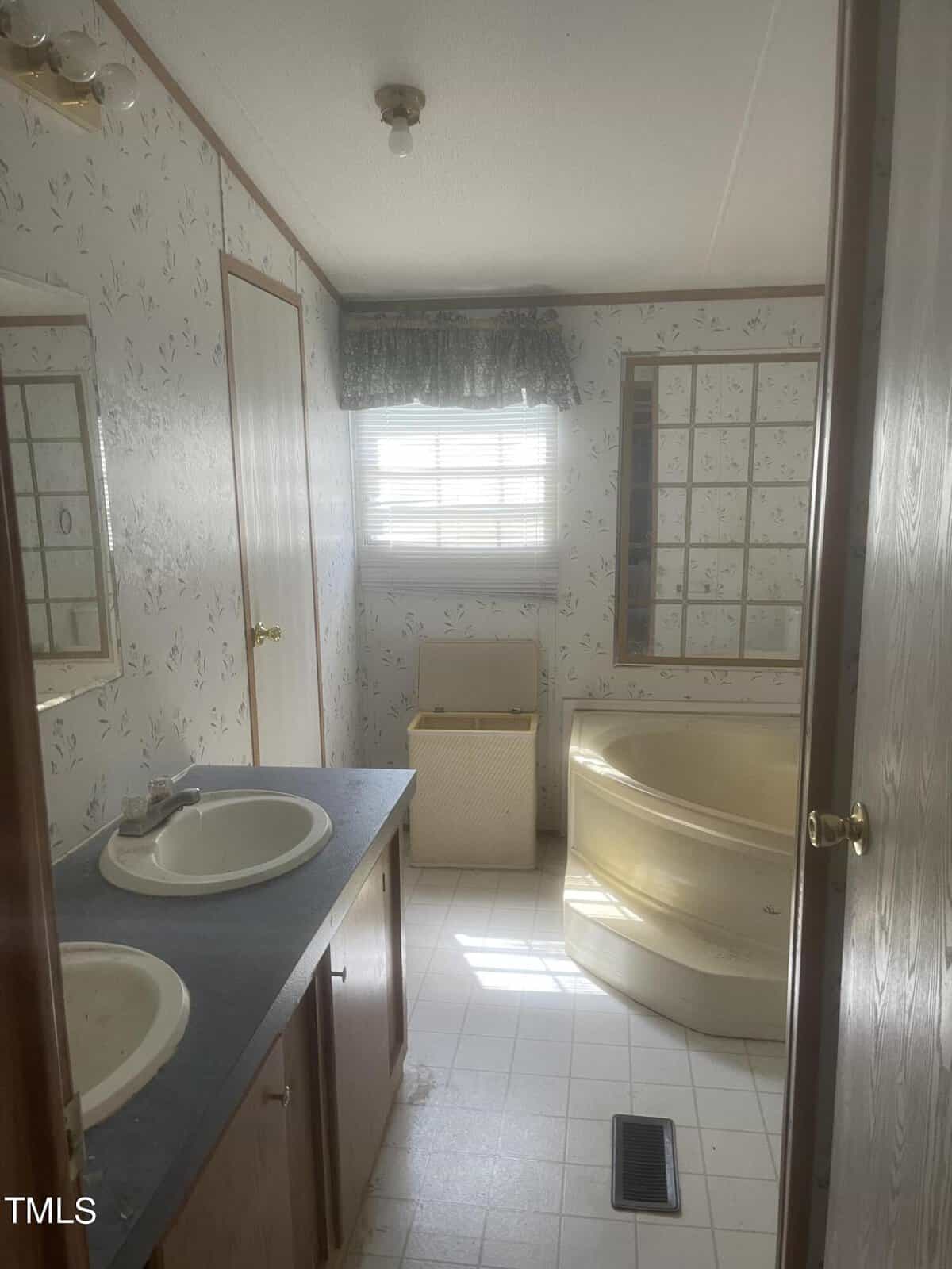 A small bathroom with floral wallpaper features a double sink vanity, a yellow corner bathtub, a window with a green valance curtain, and a laundry basket. The room has a light fixture on the ceiling and a tiled floor with a floor vent.