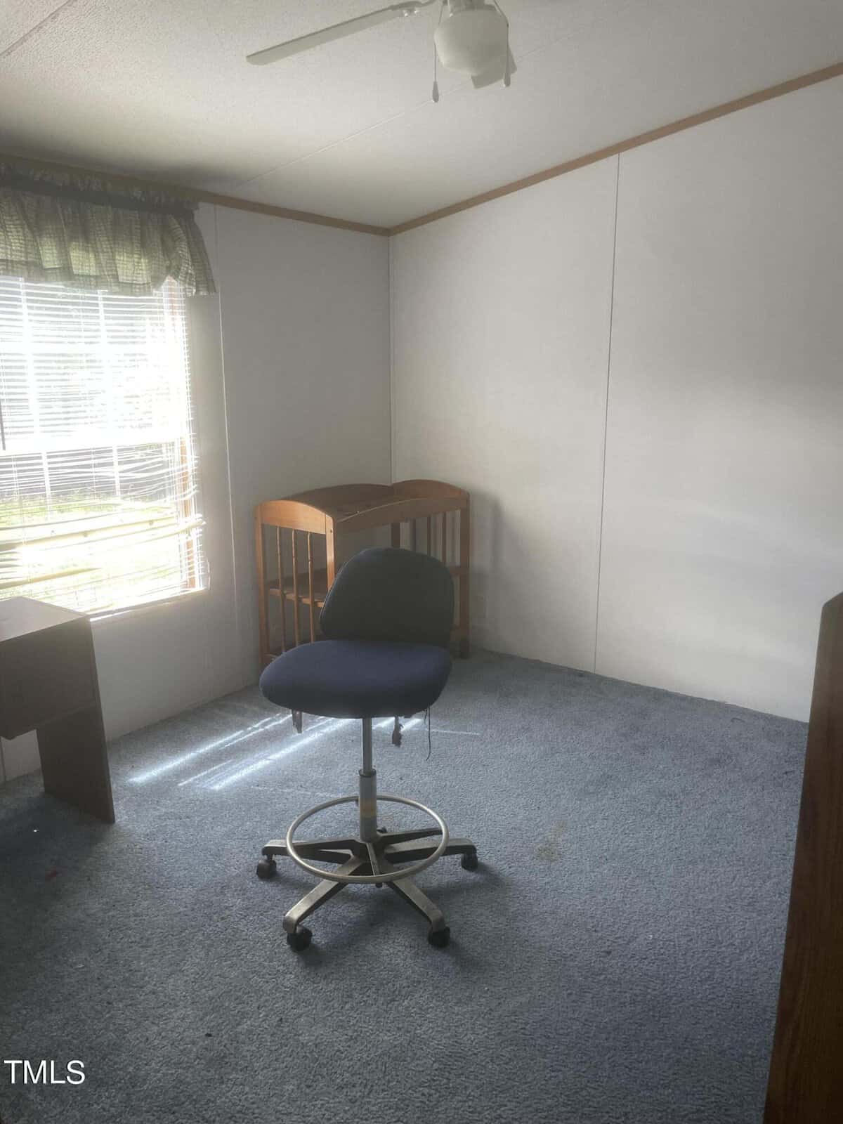 An empty room with a blue swivel chair on a blue carpet. A small wooden crib is against the wall near a window with blinds and partially open curtains. The walls are plain white, and there is a ceiling fan overhead.