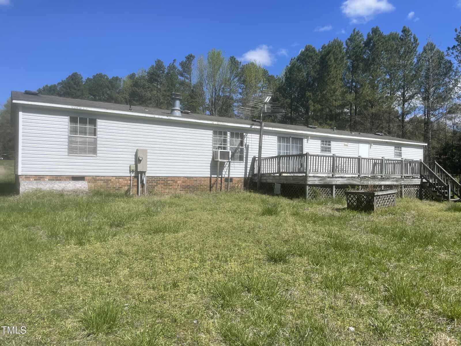 A single-story, white mobile home with a wooden deck and stairs sits in a grassy yard. Surrounded by trees under a clear blue sky, the house features multiple windows and a small chimney.