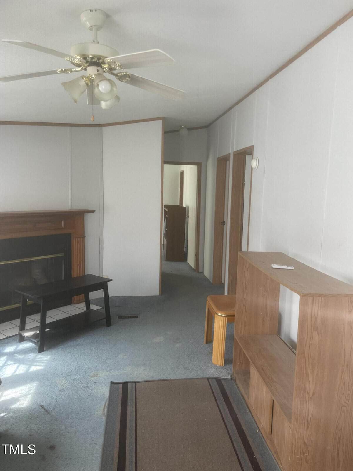 A living room with light-colored walls and blue carpet. There's a ceiling fan, a wooden fireplace with a black mantle, a small black bench, and a wooden shelf with an orange stool. A hallway is visible in the background.