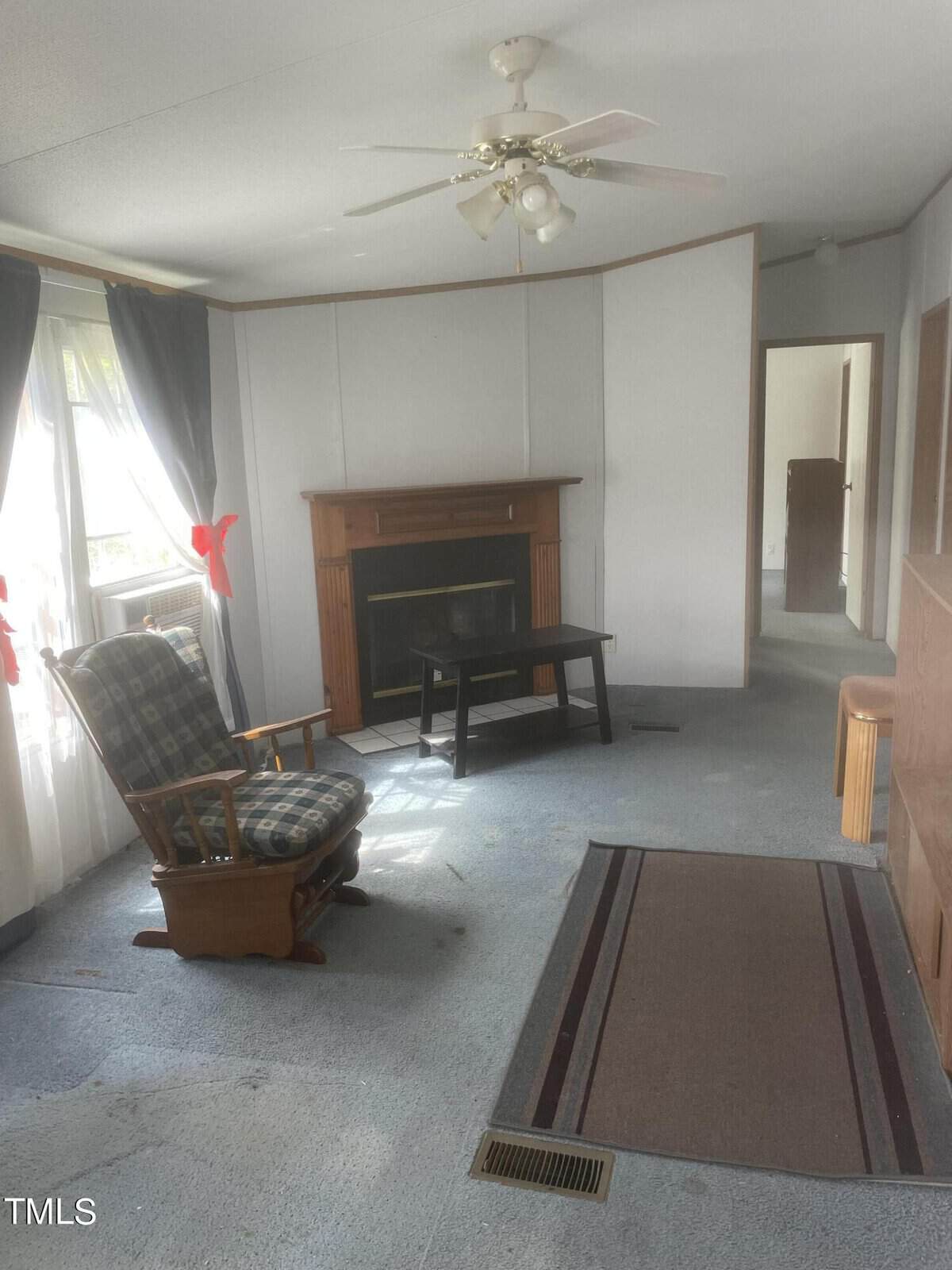 A cozy living room on Oxford's charming 2142 Mountain Creek Road features a wooden rocking chair, a small black table, and a brown fireplace. Large windows with curtains invite light into the space. A ceiling fan whirs above, and a doorway opens to another room.