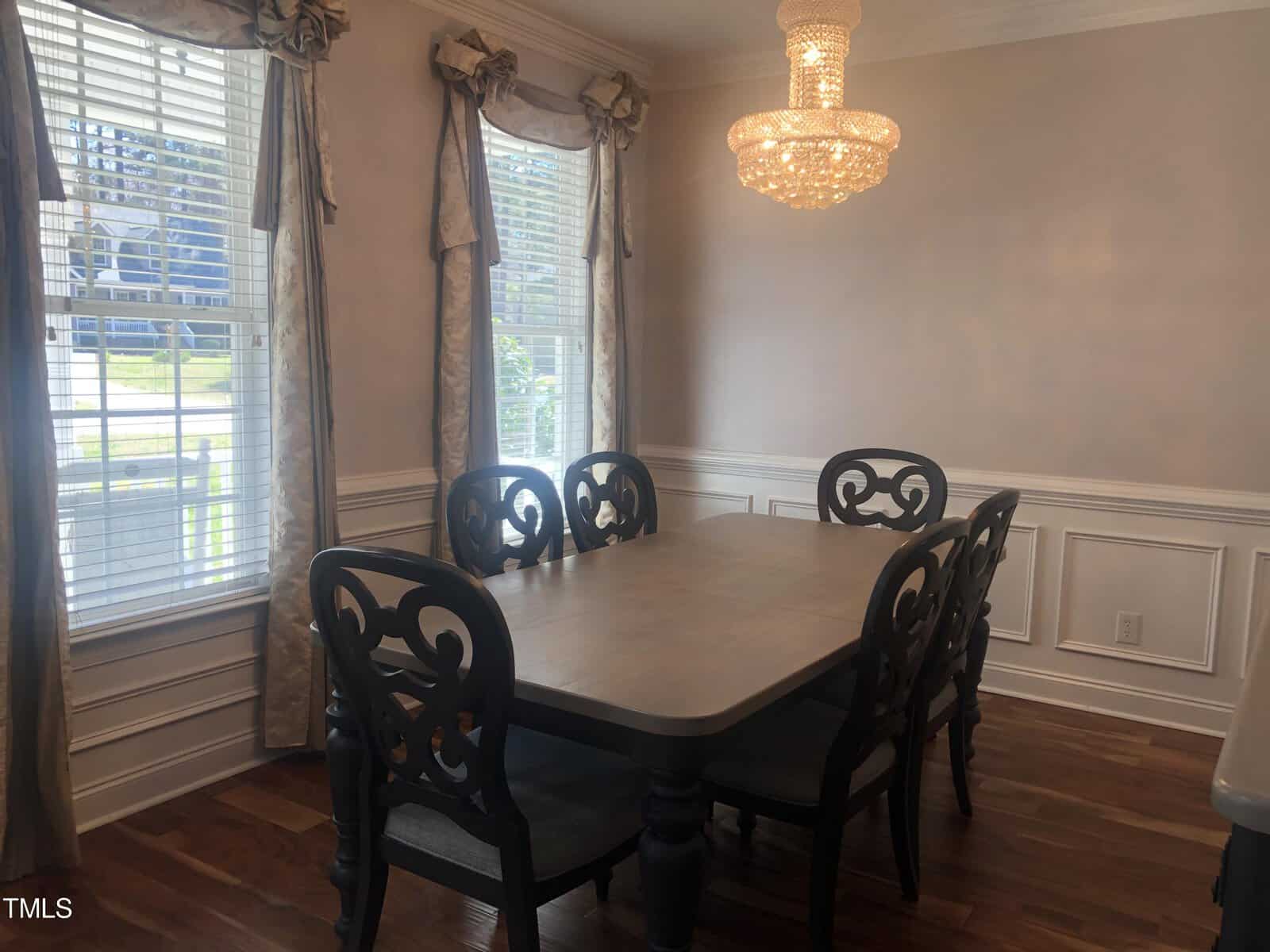 A dining room featuring a rectangular wooden table surrounded by six ornate black chairs, a crystal chandelier above, beige walls with white trim, and windows with elegant drapery.
