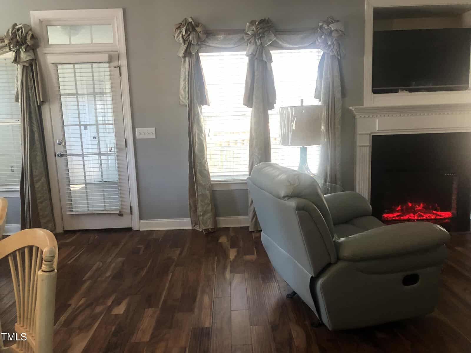 A cozy living room featuring a white fireplace with a glowing electric fire, two draped windows, a recliner, and hardwood flooring. a television is built into the wall above the fireplace.