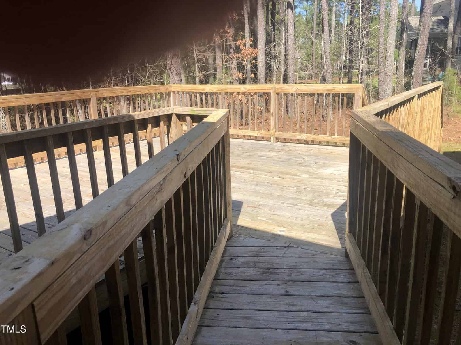A wooden deck with railings, surrounded by trees. the deck extends into a sunny, wooded area, viewed from steps leading down to it.