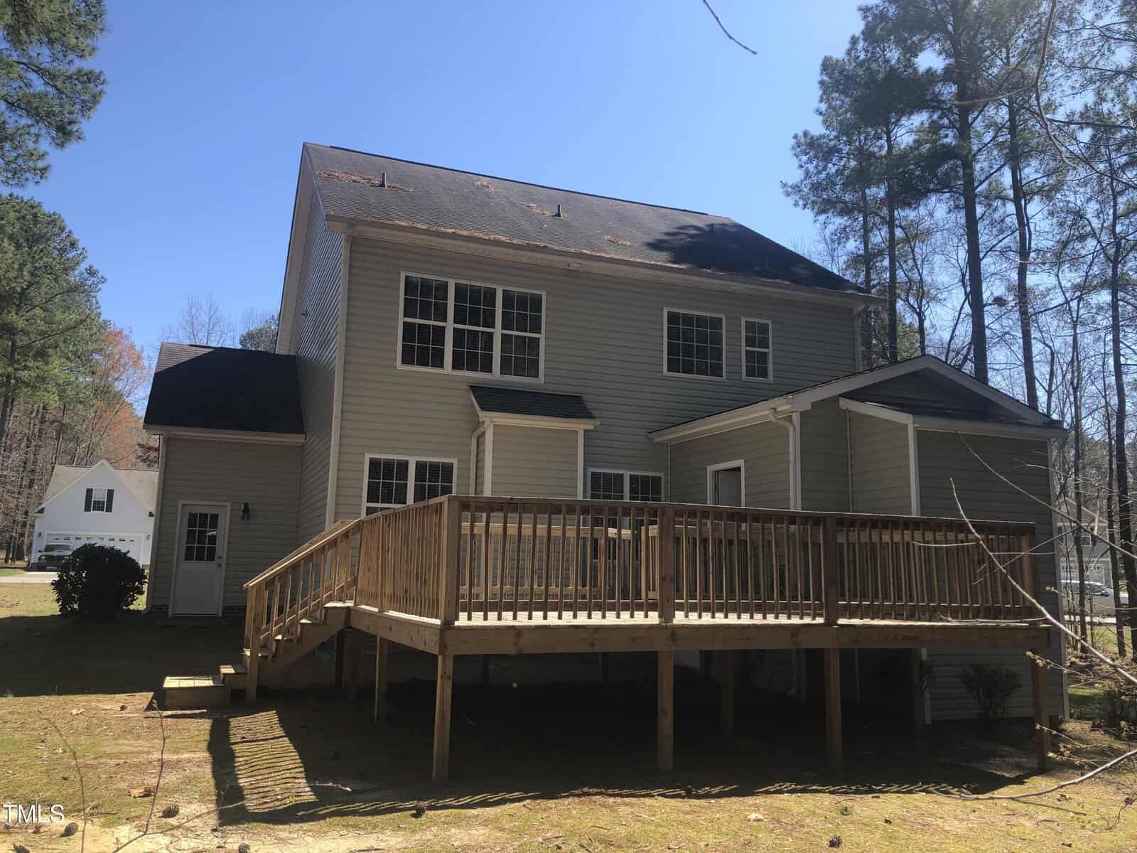 A two-story beige house with a large wooden deck, surrounded by tall pine trees on a clear sunny day. the house has black shingles and several windows, located in a suburban area.