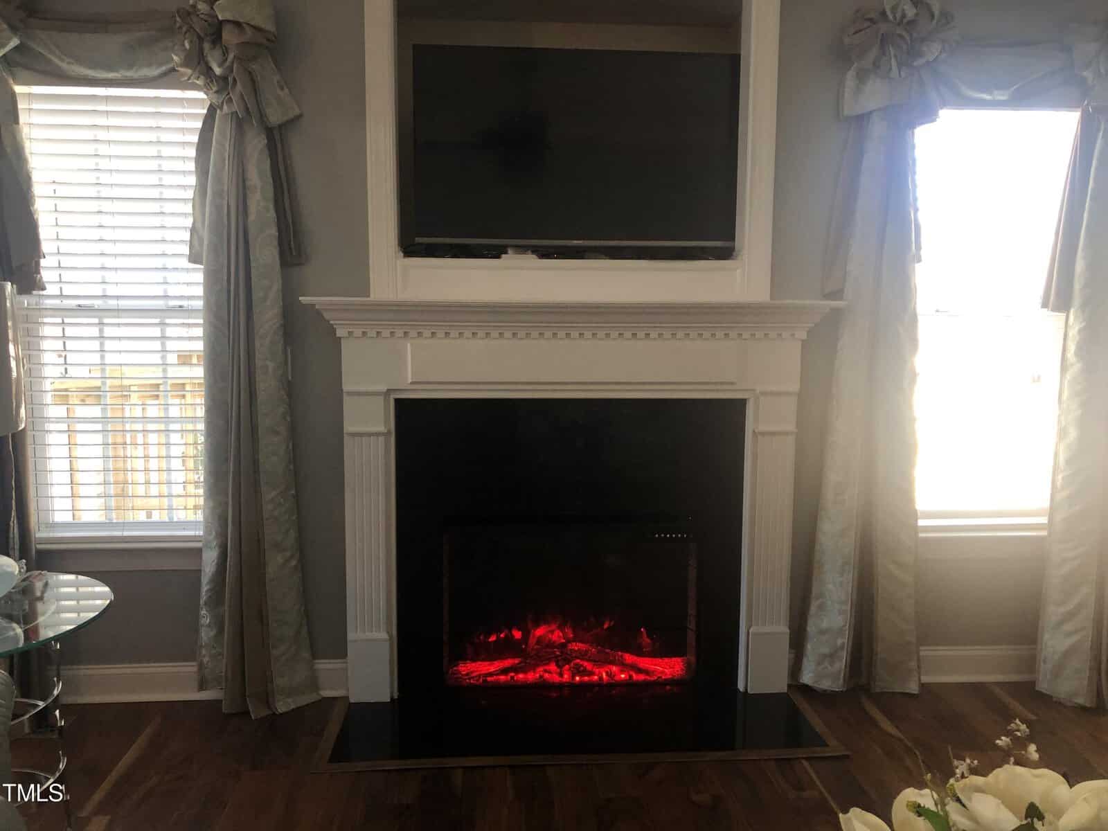 A cozy living room with an electric fireplace glowing red beneath a mounted television, flanked by two large windows draped with elegant gray curtains.