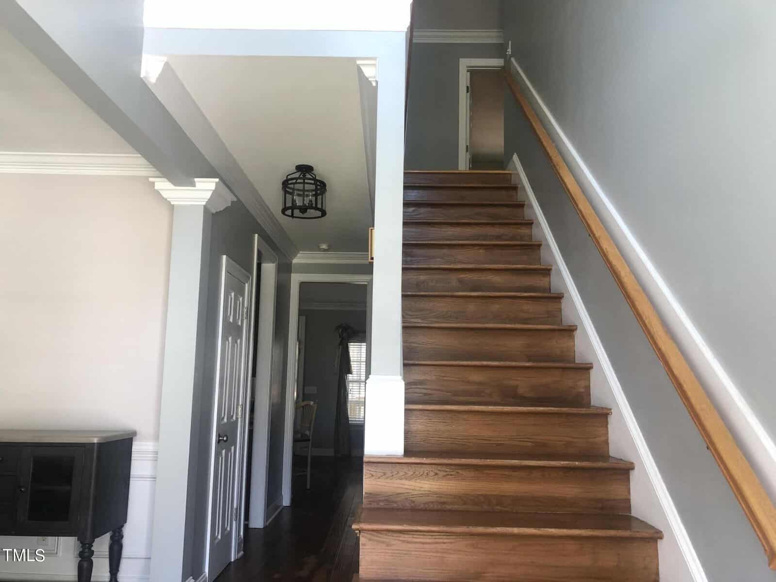 Interior of a home showing a wooden staircase with a handrail leading to the upper floor, adjacent to gray-painted walls and a corridor leading to other rooms.