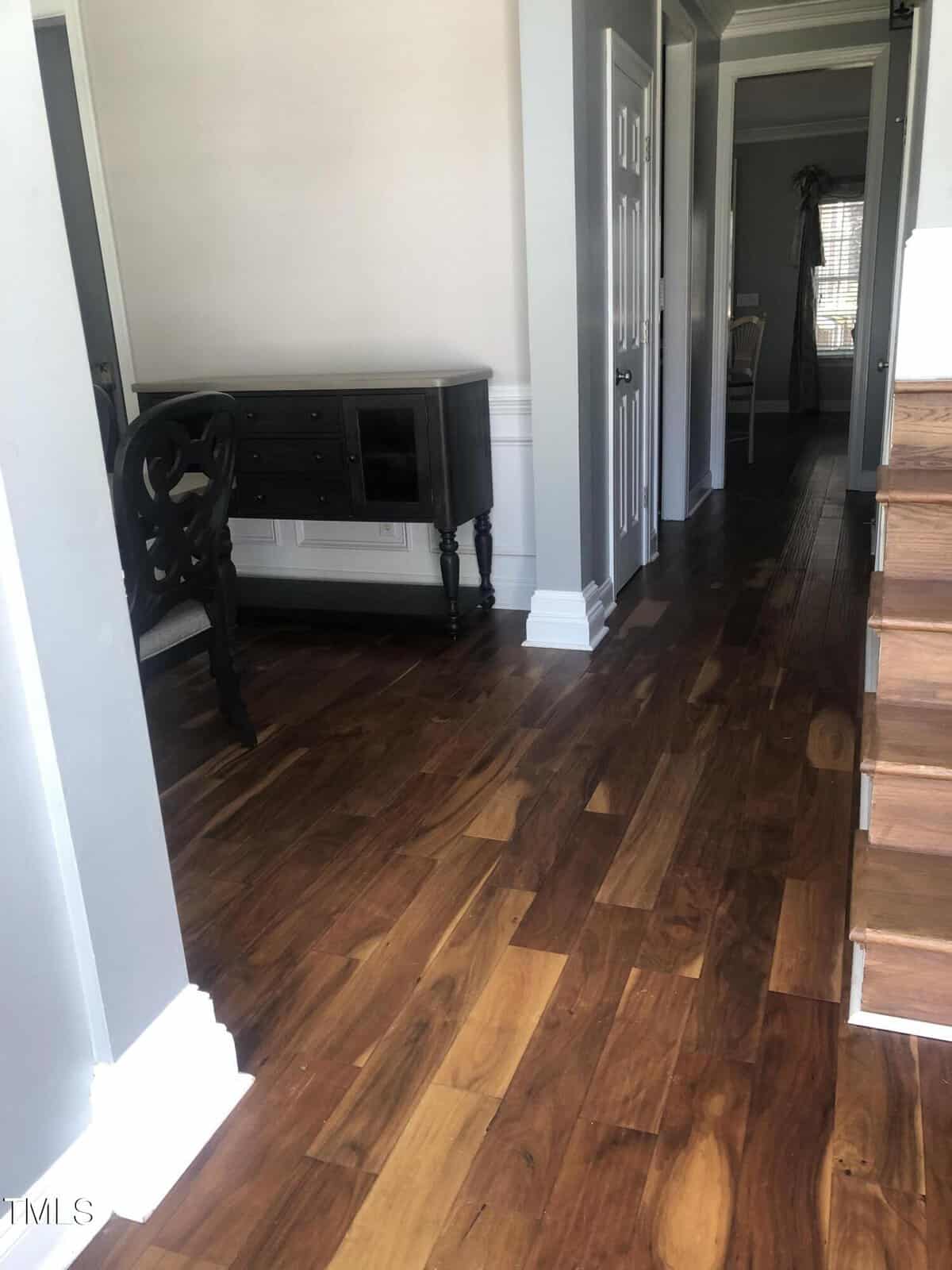 A hallway featuring shiny, varied-tone wood flooring, with a black cabinet and a chair on the right side. doorways branch off from the hallway leading to other parts of the house.