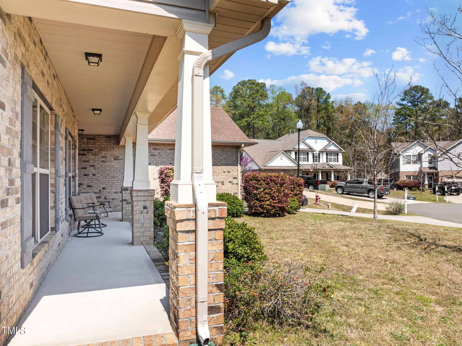 Front porch of a suburban home with light brown brick, featuring a small seating area. The view includes a grassy yard and neighboring houses on a sunny day with a few clouds in the sky.