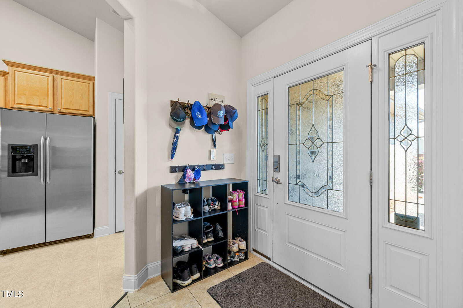 Entryway with a glass-paneled front door and sidelights. A shelf holds various shoes, and above it, hooks carry several caps and a small bag. A stainless steel refrigerator is visible in an adjacent kitchen area. A doormat lies on the floor.