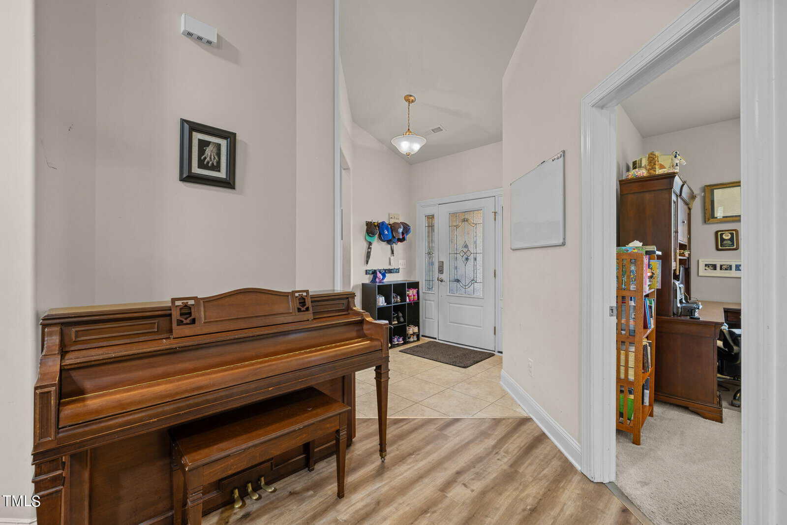 A cozy hallway with a wooden piano on the left, a door with a glass panel ahead, and a glimpse of an office on the right. The hallway features light-colored walls, wooden flooring, and a small chandelier.