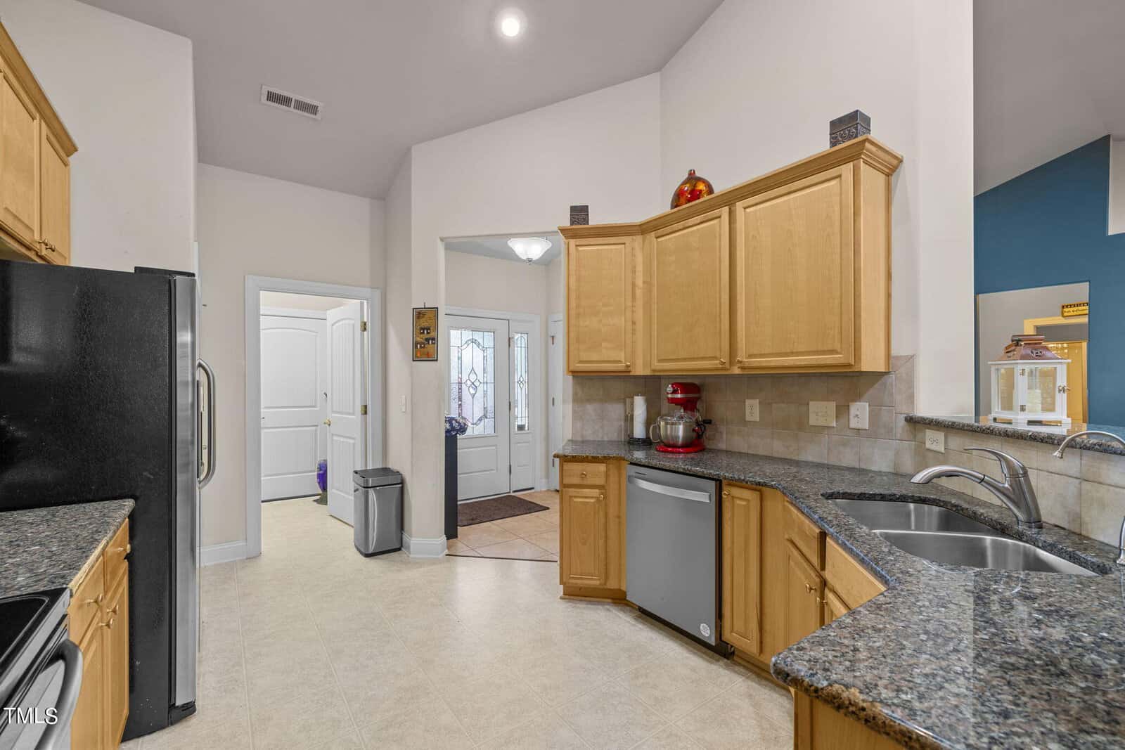 A bright, modern kitchen featuring granite countertops, light wood cabinets, stainless steel appliances, and a double sink. A red mixer sits on the counter. The kitchen opens to an entryway with a decorative glass door and a tiled floor.