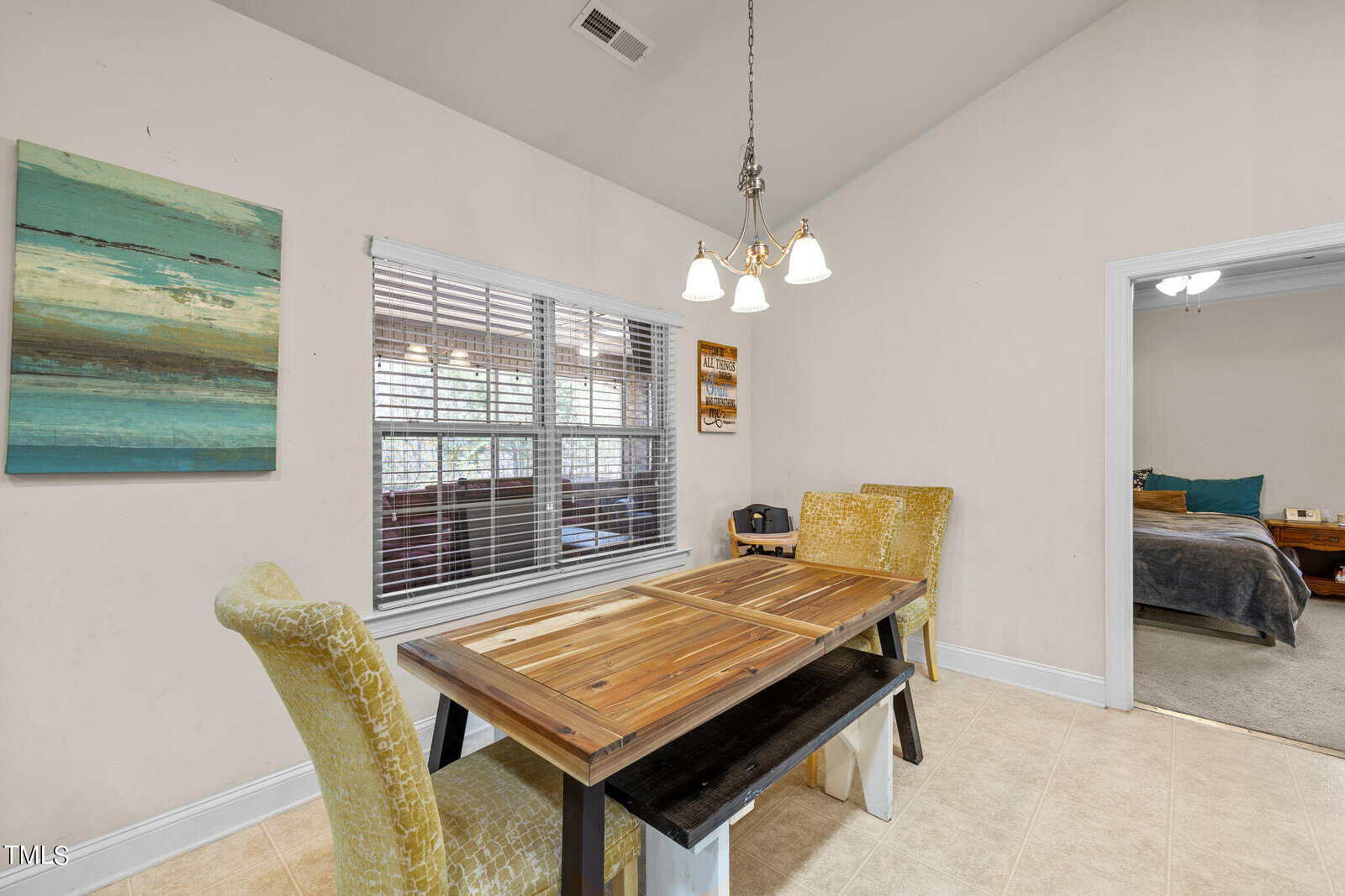 A dining area with a wooden table and two cushioned chairs. A painting hangs on the light-colored wall. A window with blinds is to the left, and a doorway leads to a bedroom with a bed and nightstand visible. A ceiling light fixture is above.