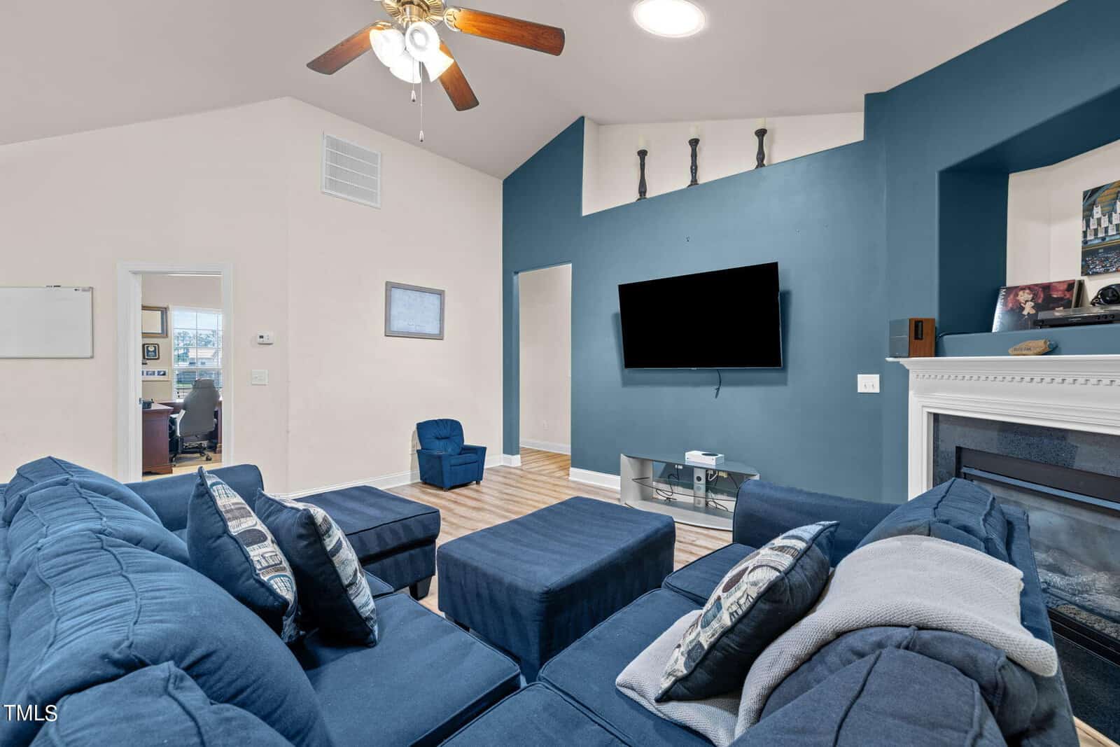 A cozy living room with a large blue sectional sofa facing a flat-screen TV mounted on a teal accent wall. There's a ceiling fan above, a fireplace on the right, and an open doorway leading to a well-lit room. Natural light fills the space.