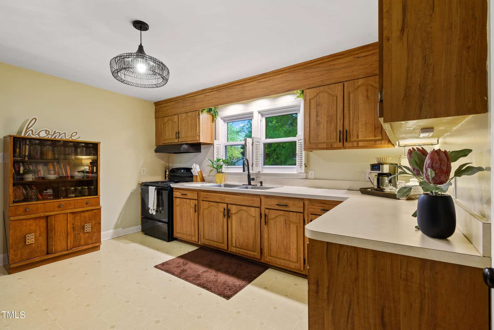 A cozy kitchen with wooden cabinets, a white countertop, and a small rug. A window with open shutters lets in natural light. A black oven and fridge are on the left. Decorative elements include a shelf labeled "home" and a vase with flowers.