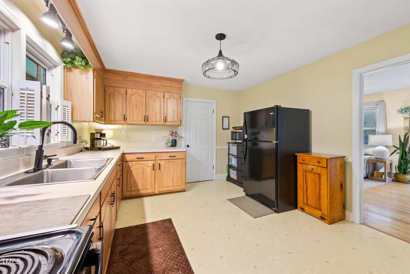 A spacious kitchen with wooden cabinets, a black refrigerator, and a double sink. There's a door leading to another room. The floor is tiled, and the walls are painted light yellow. A pendant light hangs from the ceiling.