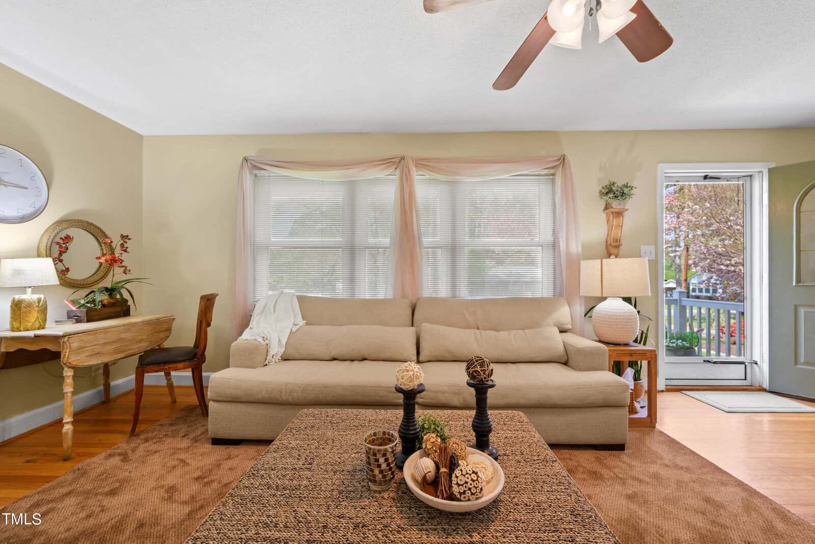 A cozy living room featuring a beige sofa with pillows, a wooden coffee table with decorative items, and a patterned rug. A small desk with a chair and a lamp sits to the left. The room has a ceiling fan and large windows letting in natural light.