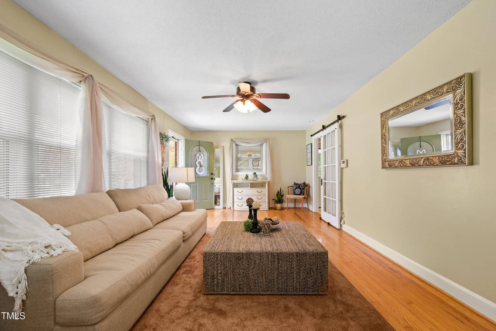 A cozy living room with a beige sofa, a large woven coffee table, and a ceiling fan. French doors lead to a decorated cabinet and mirror. Wooden floors and light-colored walls create a warm ambiance. Large windows bring in natural light.