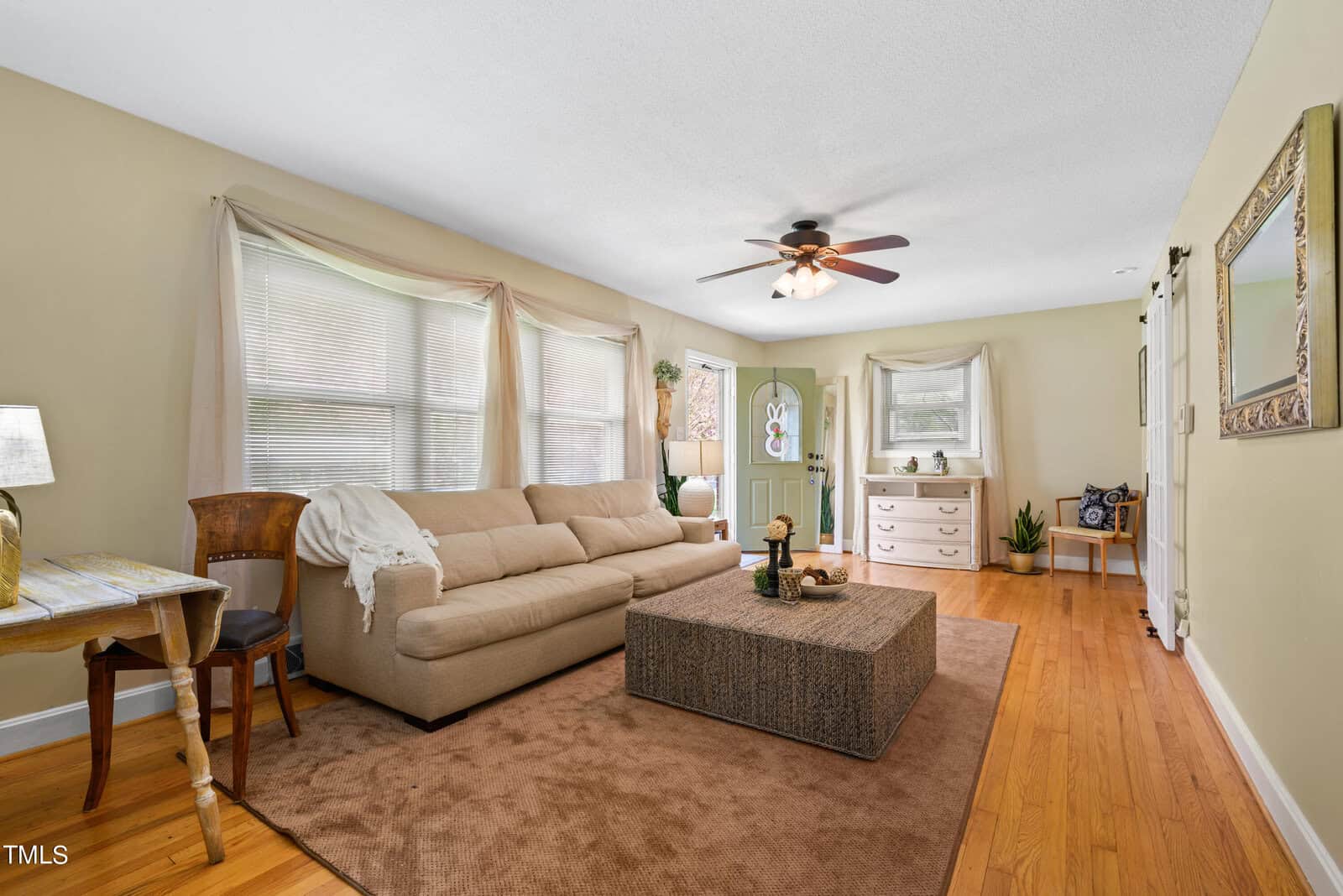 A cozy living room with beige walls and hardwood floors. There's a large beige sofa, a wicker coffee table, and a ceiling fan overhead. Large windows with curtains illuminate the space, and decorative items are placed throughout.