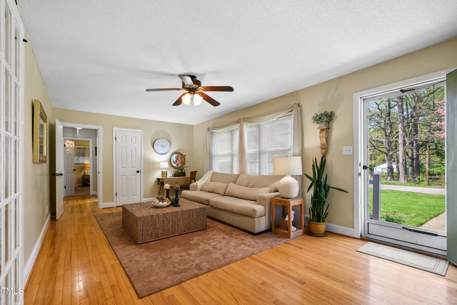 A cozy living room features a beige sofa, a woven coffee table, and large windows with light curtains. A ceiling fan hangs above, and there's a door leading outside. Wooden floors and potted plants add warmth and charm to the space.
