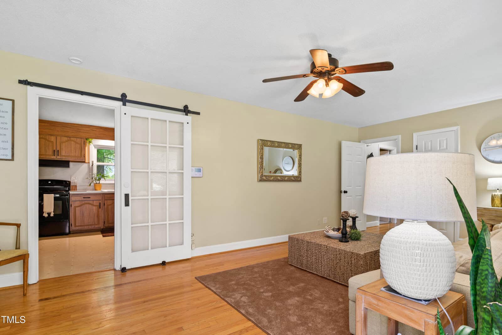 A cozy living room with hardwood floors, a ceiling fan, and a large beige sofa. A white sliding barn door leads to a kitchen. A large lamp and green plant decorate a side table. A mirror hangs on a light green wall.