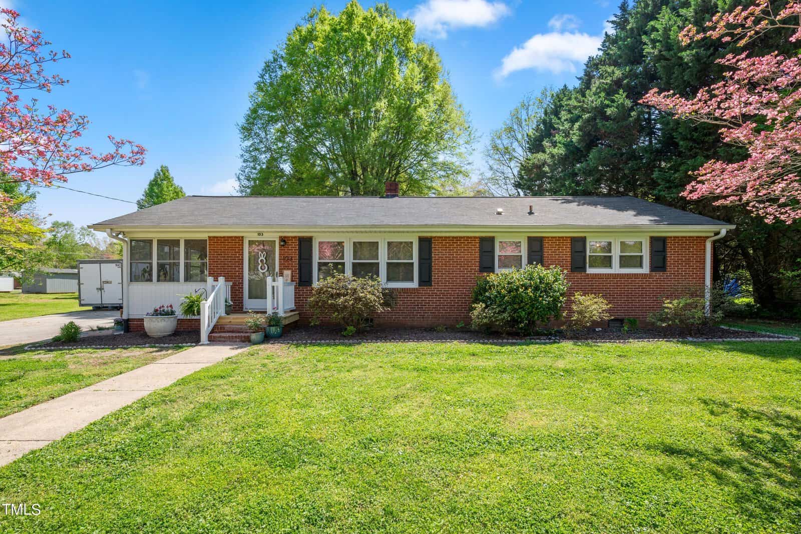 Located at 103 Pine Tree Drive, this charming brick ranch-style house in Oxford boasts a gray roof and is surrounded by lush greenery. Large windows grace the facade, while the small front porch with white railings invites you in along a pathway lined with blooming pink flowers and shrubs.