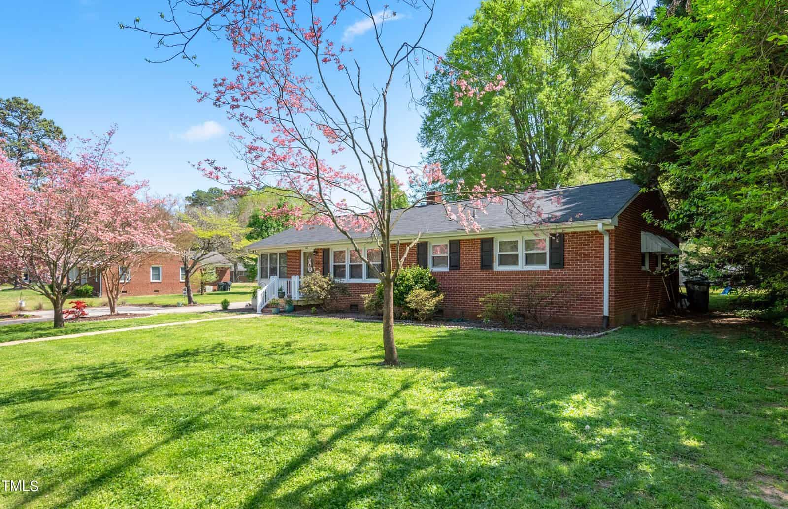 A charming red-brick house surrounded by lush green grass and blooming pink trees under a bright blue sky. The home has white-framed windows and a small front porch, creating a welcoming and serene atmosphere.