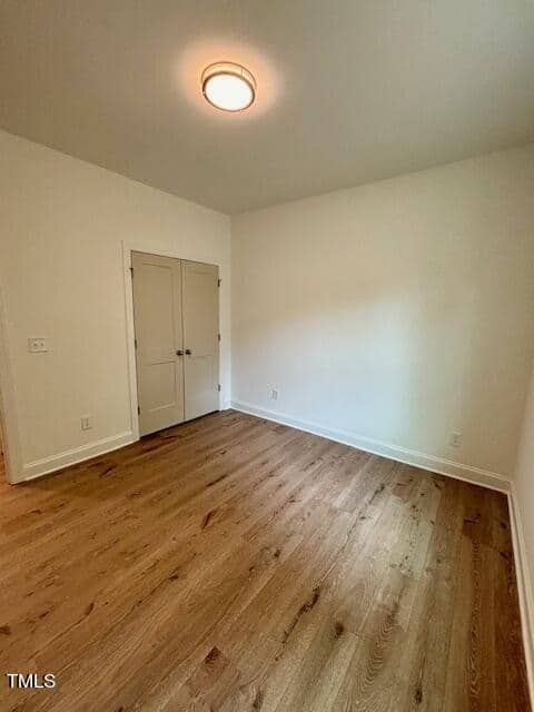 A simple, empty room with white walls and hardwood flooring featuring a closed white door and a ceiling light.