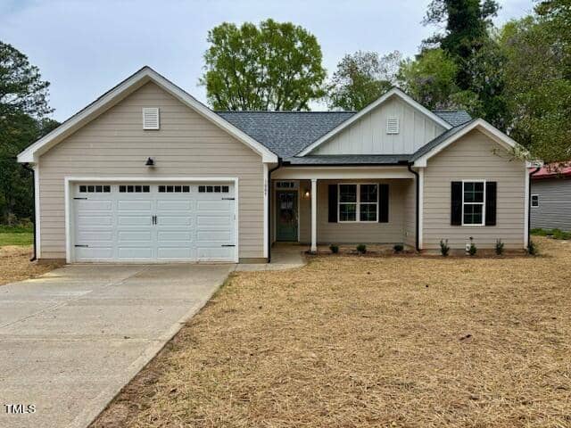 A new single-story house in Henderson, NC, with a gray siding exterior, featuring a large white garage door and a covered entrance with a gabled roof, set against a backdrop of trees.
