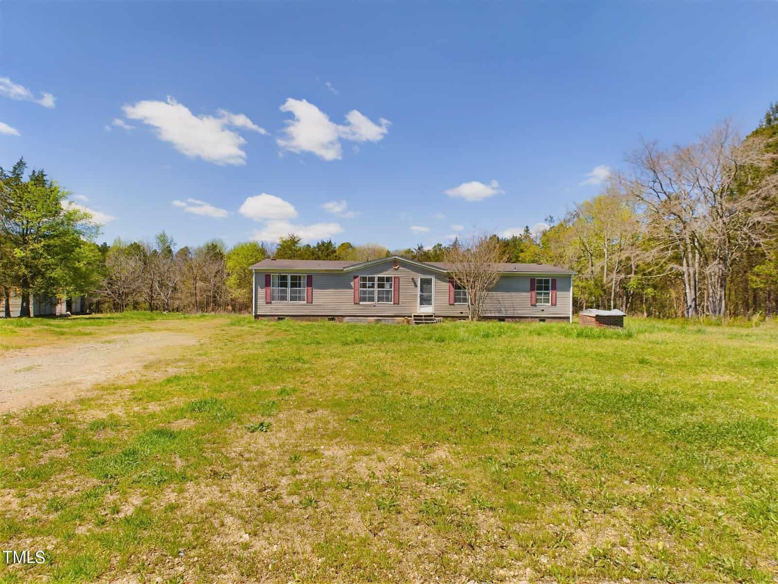 A single-story house with light-colored siding sits on a large grassy lot under a bright blue sky with scattered clouds at 9515 NC 49. Located in Roxboro, the property features sparse trees in the background, offering a serene and picturesque setting.