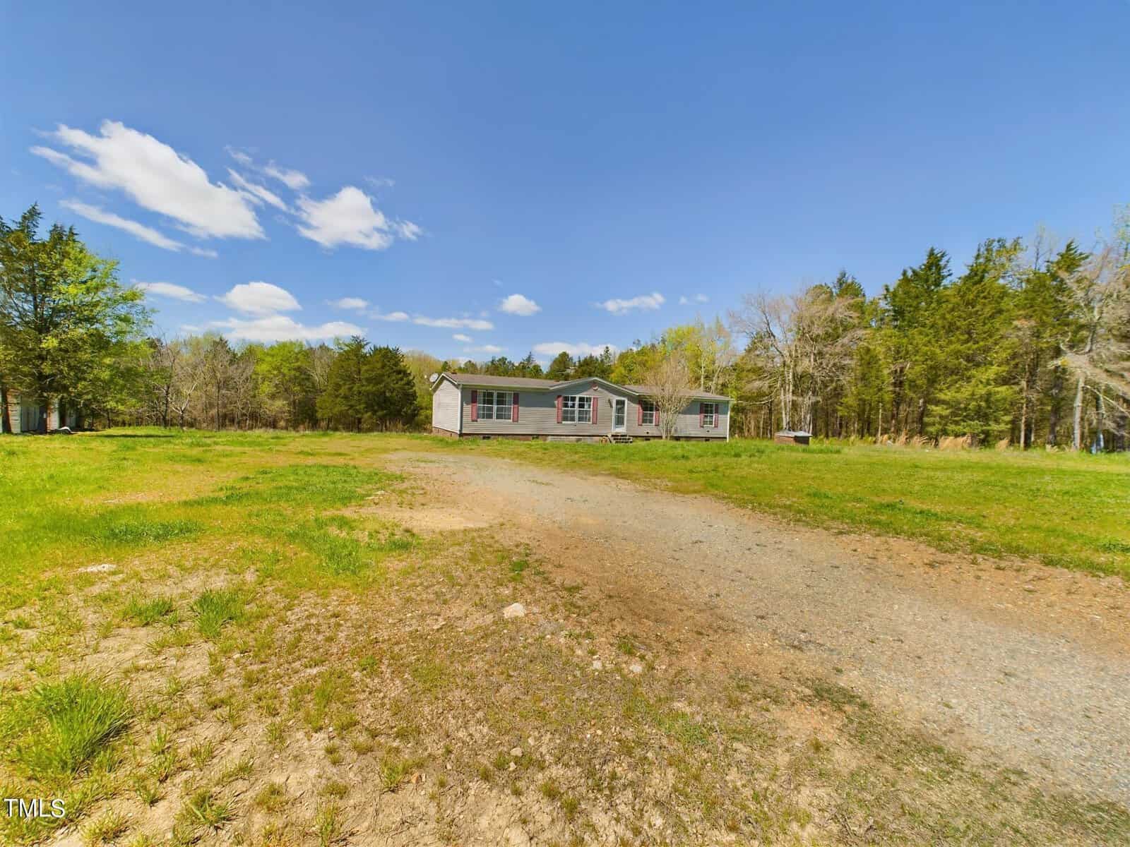 A rural landscape with a single-story house surrounded by a grassy yard and a dirt driveway. The sky is bright blue with scattered clouds, and trees border the property.